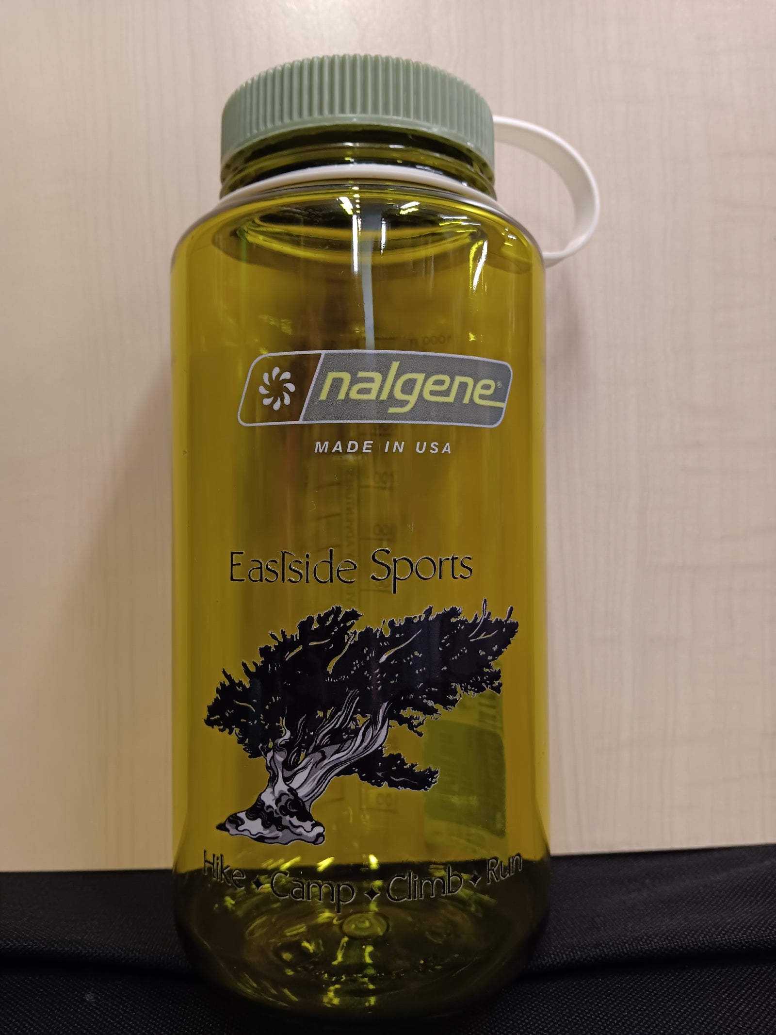 A translucent yellow-green Nalgene water bottle with a green cap and white carry loop, featuring Eastside Sports branding and a black tree graphic on a beige wall background.