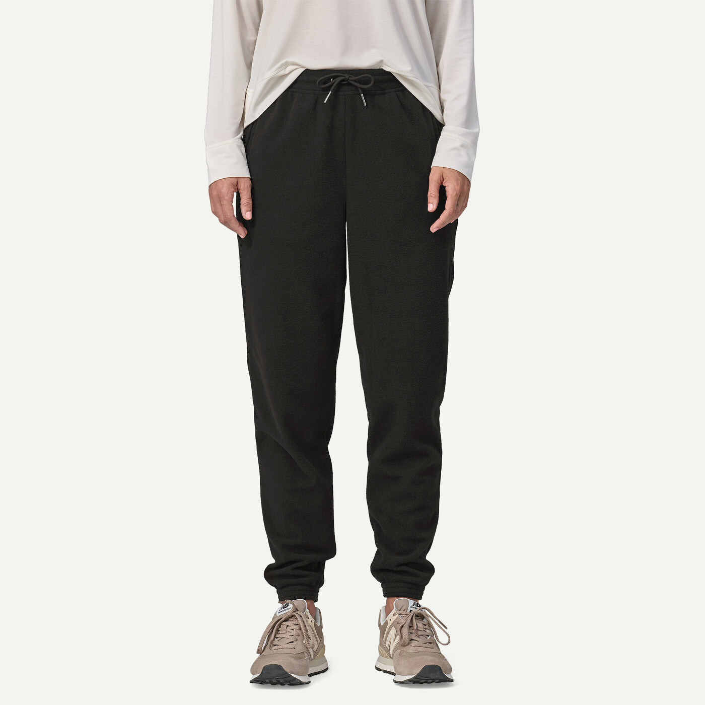A person wearing black jogger sweatpants with a drawstring waist, paired with a light beige long-sleeve top and beige sneakers, shown against a plain light gray studio background.