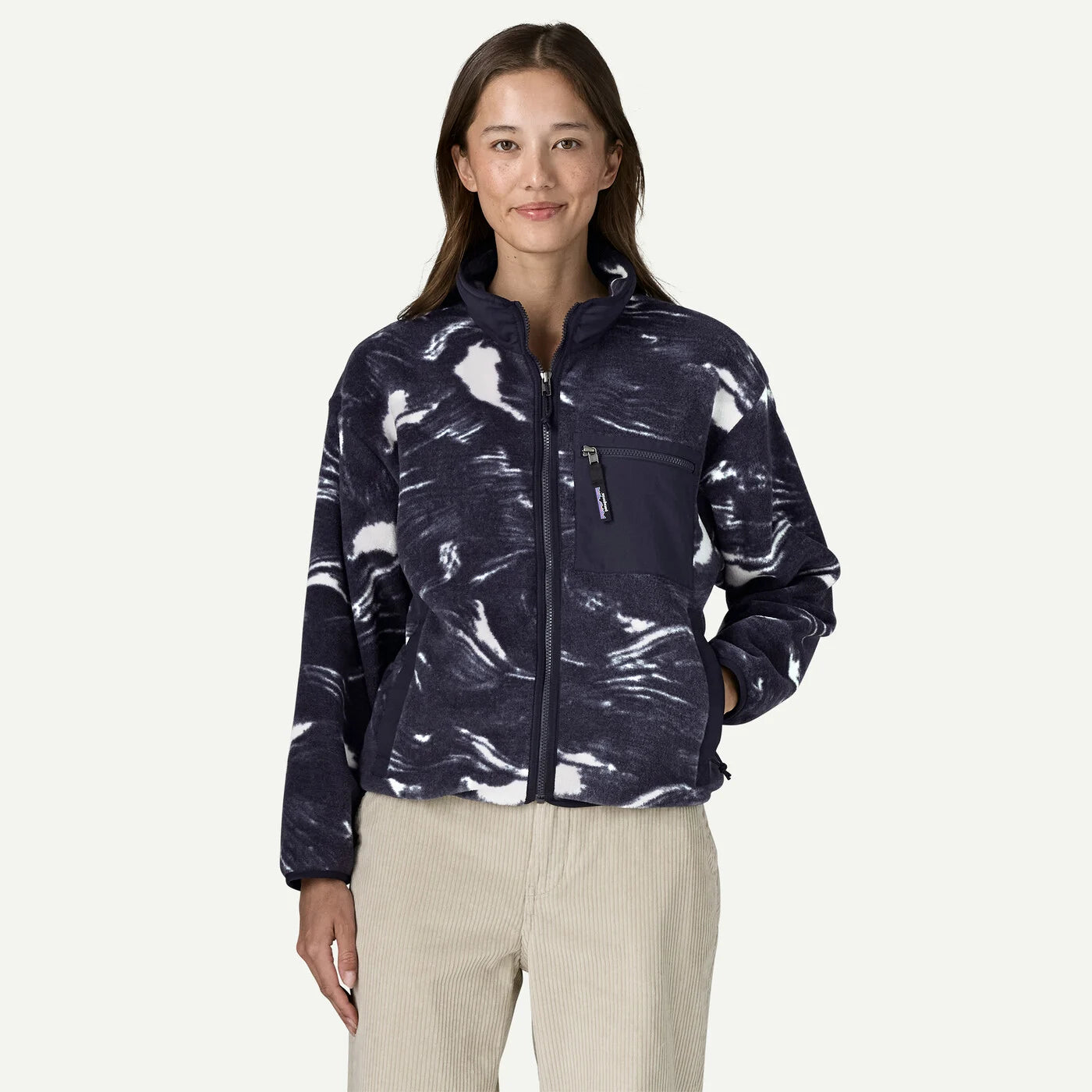 A woman modeling a navy blue fleece jacket with a white abstract marbled pattern, paired with beige pants, against a solid off-white background.