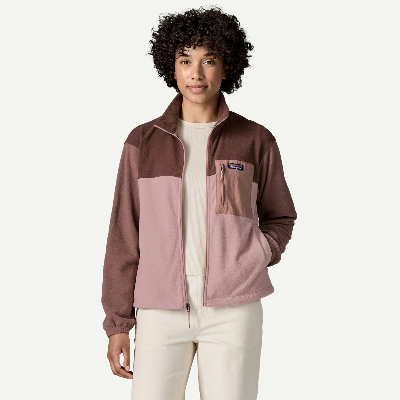 A woman modeling a Patagonia fleece jacket with a colorblock design in dark brown and dusty pink, worn over a cream top and cream pants against a light off-white background.