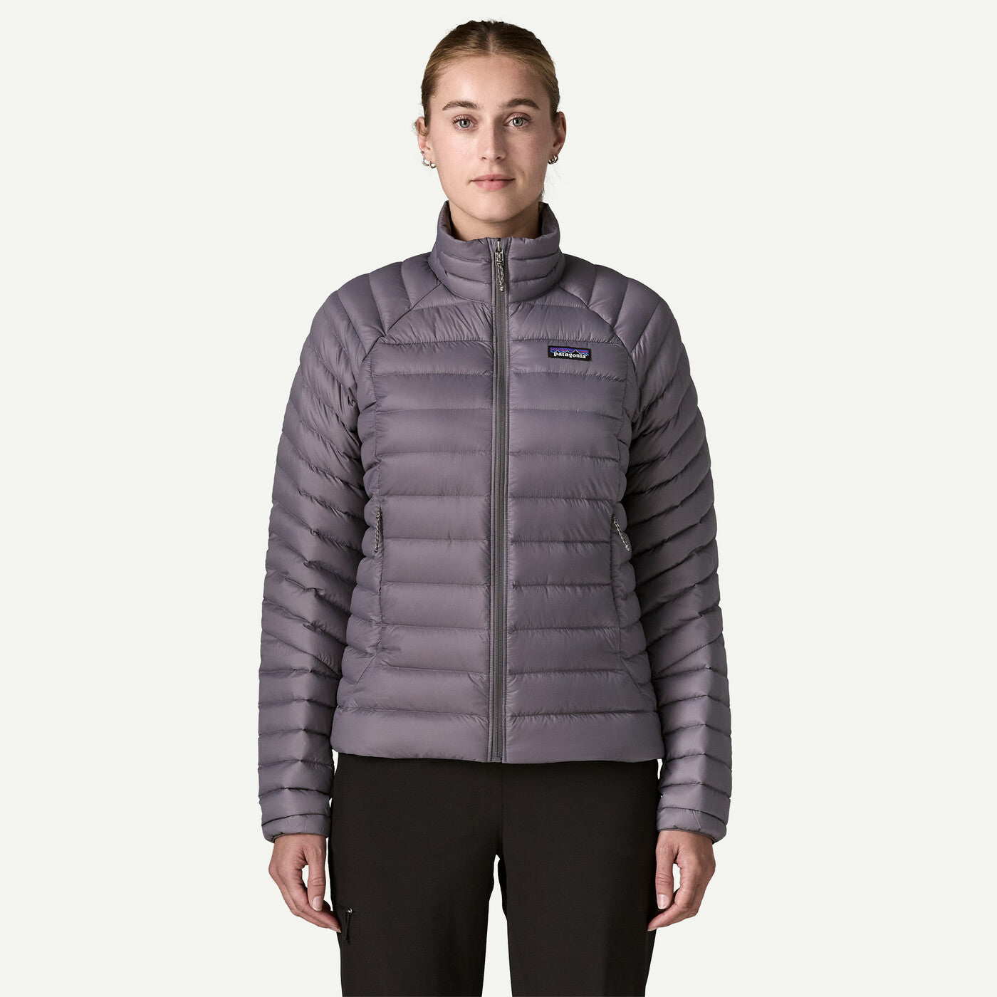 A woman modeling a Patagonia women's quilted puffer jacket in a muted grayish purple color, paired with black pants, against a solid off-white background.
