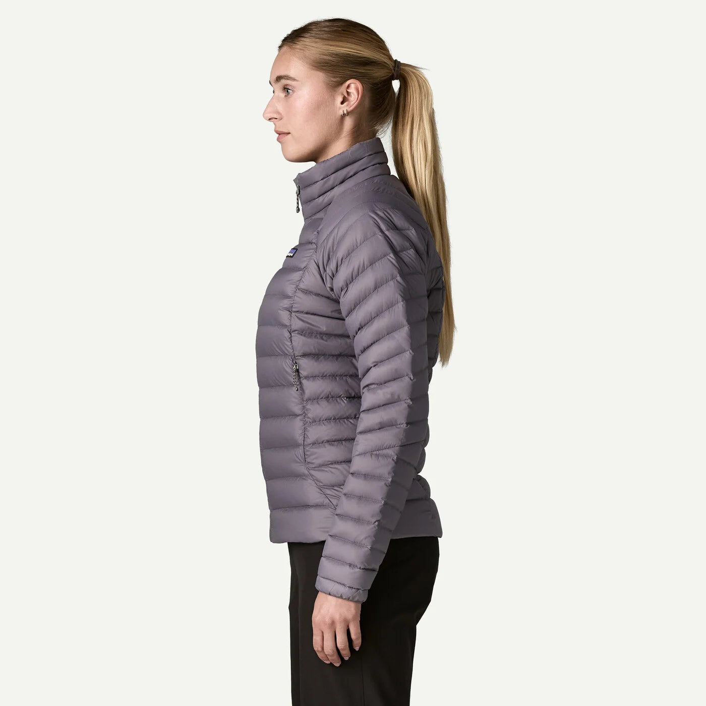 A woman in profile wearing a light gray quilted puffer jacket and black pants, standing against a solid off-white background.