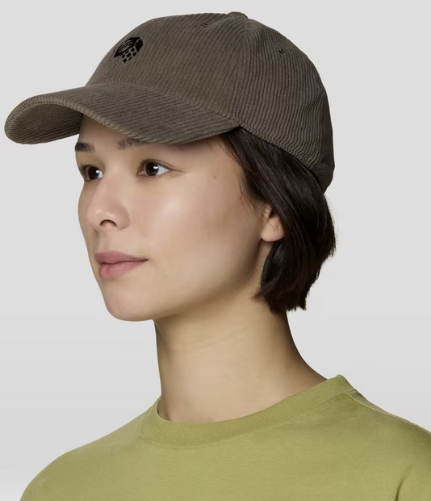 A woman in profile wearing a brown corduroy baseball cap with a small black embroidered logo on the front, paired with an olive green crewneck shirt, against a light gray studio background.
