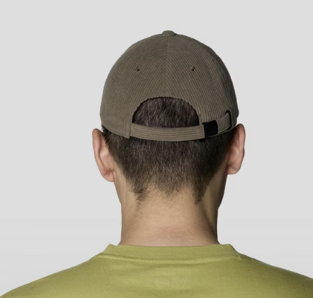 Rear view of a man wearing a corduroy baseball cap in a muted olive-brown color, paired with an olive-green t-shirt, against a light gray studio background.