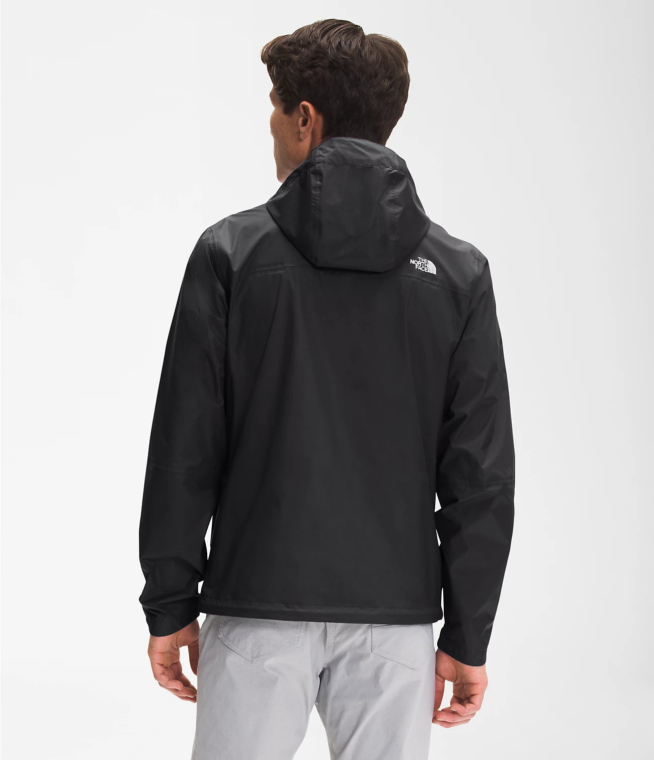 A man viewed from the back wearing a black hooded jacket with a small white The North Face logo on the upper back, paired with light gray pants, against a plain light gray studio background.