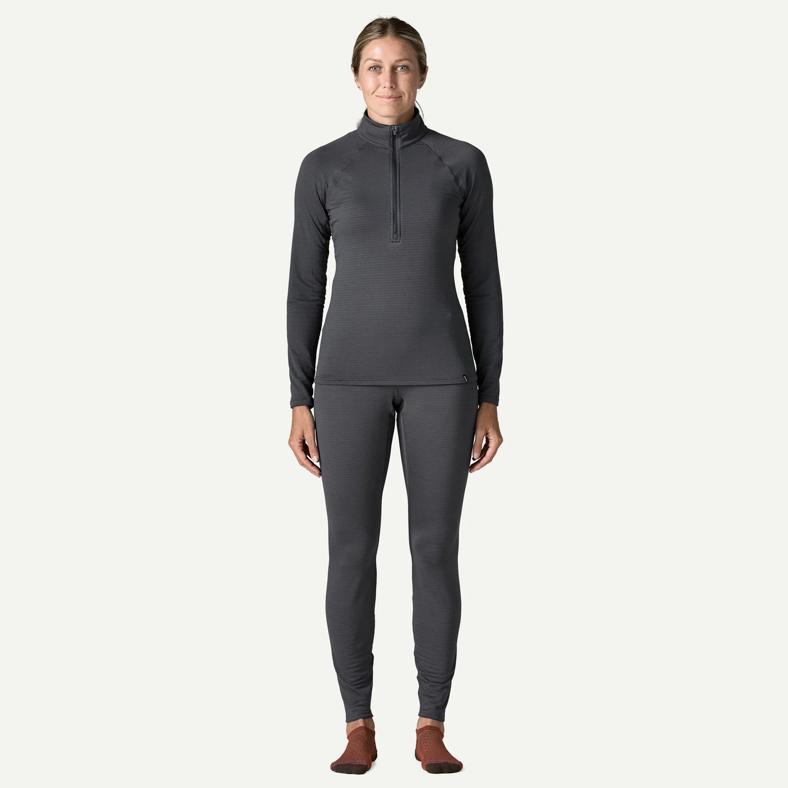 A woman modeling a dark gray long-sleeve quarter-zip top and matching full-length leggings against a light gray studio background.
