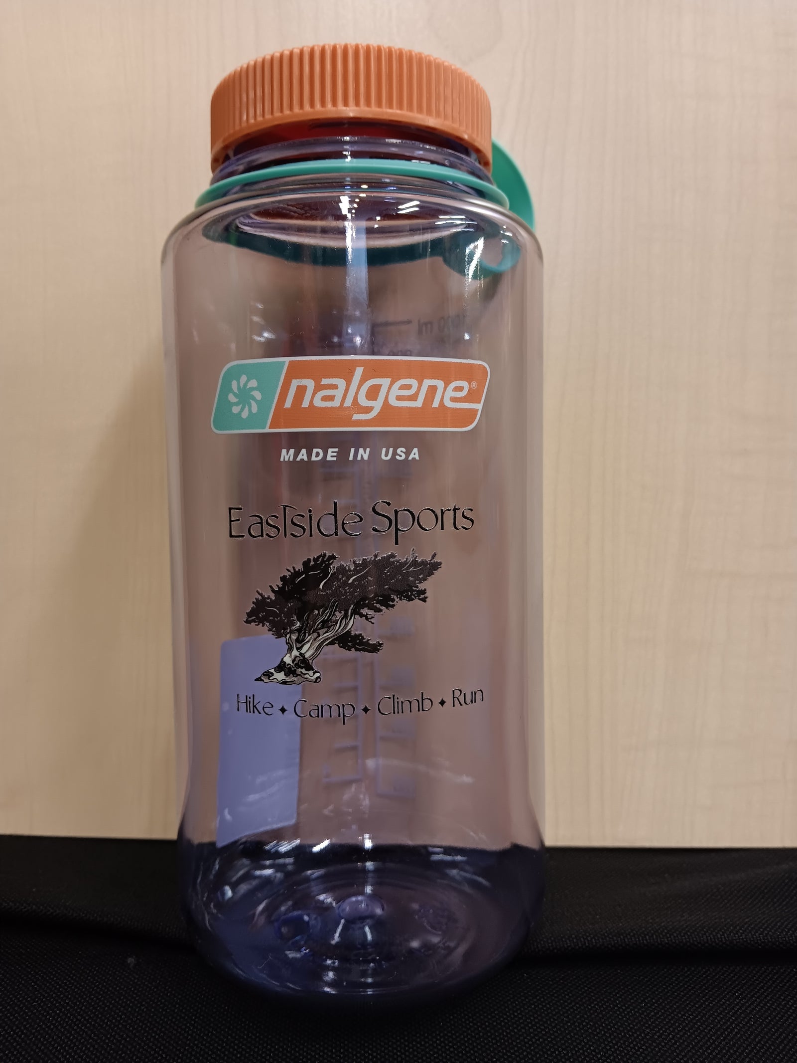 A clear plastic water bottle with an orange ribbed cap and a teal flip-straw lid, featuring the 'nalgene' and 'Eastside Sports' branding on the front, set against a light wood panel background.
