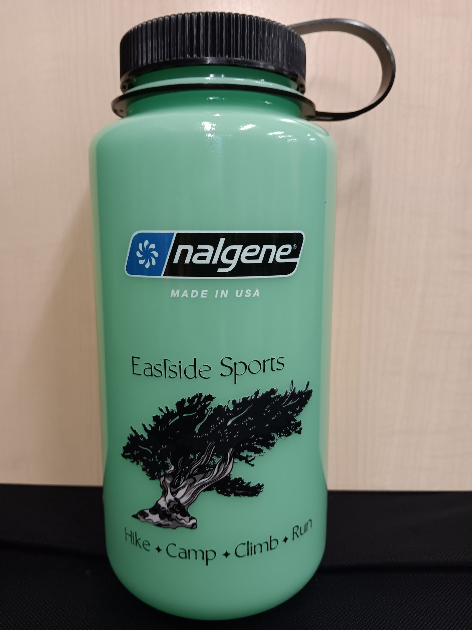 A mint green Nalgene water bottle with a black screw cap and loop handle, featuring the 'Eastside Sports' logo and a black tree graphic on the front, set against a beige wall background.
