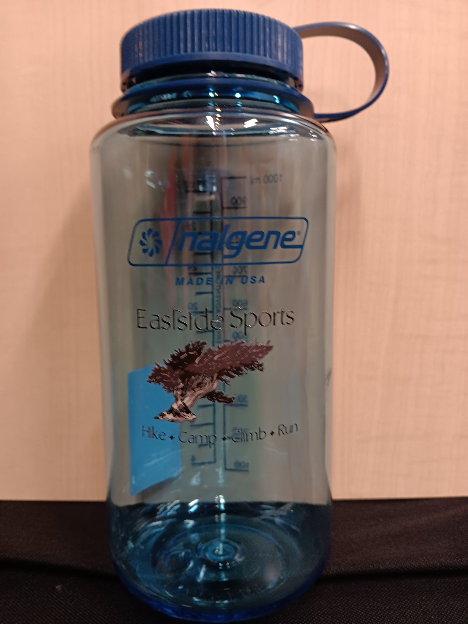A translucent blue Nalgene water bottle with a blue screw cap and integrated loop handle, standing against a beige wall.