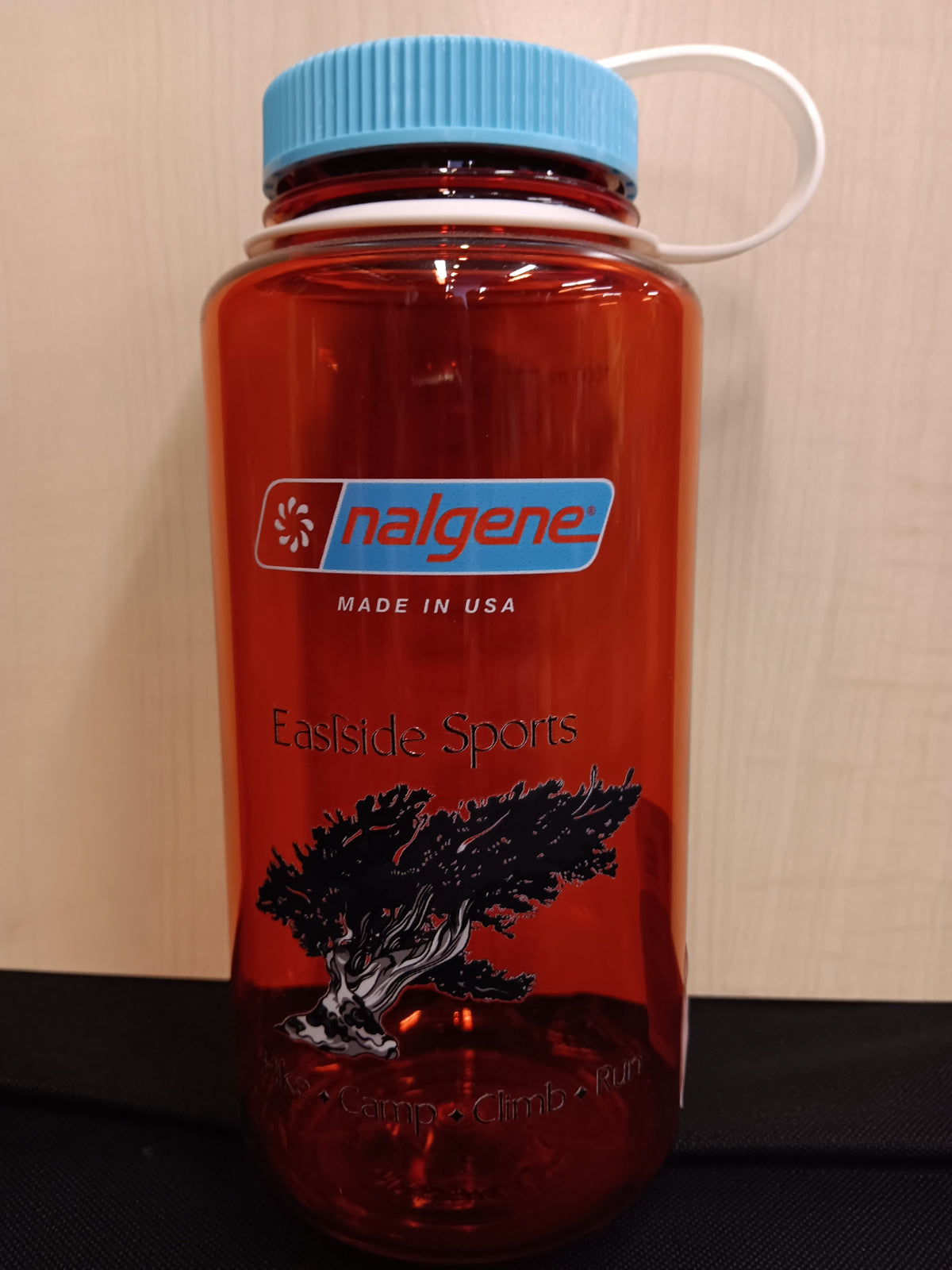 A translucent orange-red Nalgene water bottle with a light blue screw cap and white carry loop, featuring a black tree graphic and 'Eastside Sports' branding on the front.