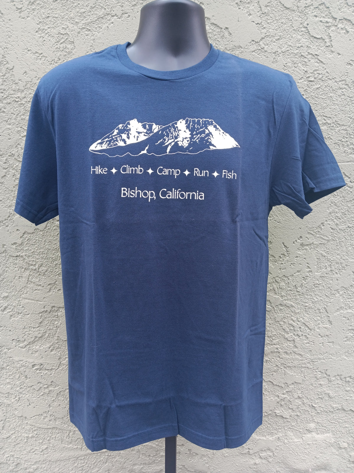 A blue short-sleeve t-shirt displayed on a dark gray mannequin torso against a textured stucco wall. The shirt features a white graphic of a mountain range with the words 'Hike + Climb + Camp + Run + Fish' and 'Bishop, California' printed below.