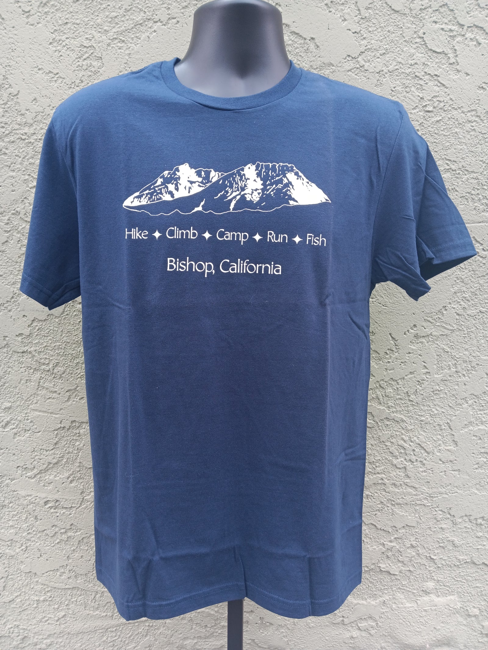 A blue short-sleeve t-shirt displayed on a dark gray mannequin torso against a textured stucco wall. The shirt features a white graphic of a mountain range with the words 'Hike + Climb + Camp + Run + Fish' and 'Bishop, California' printed below.