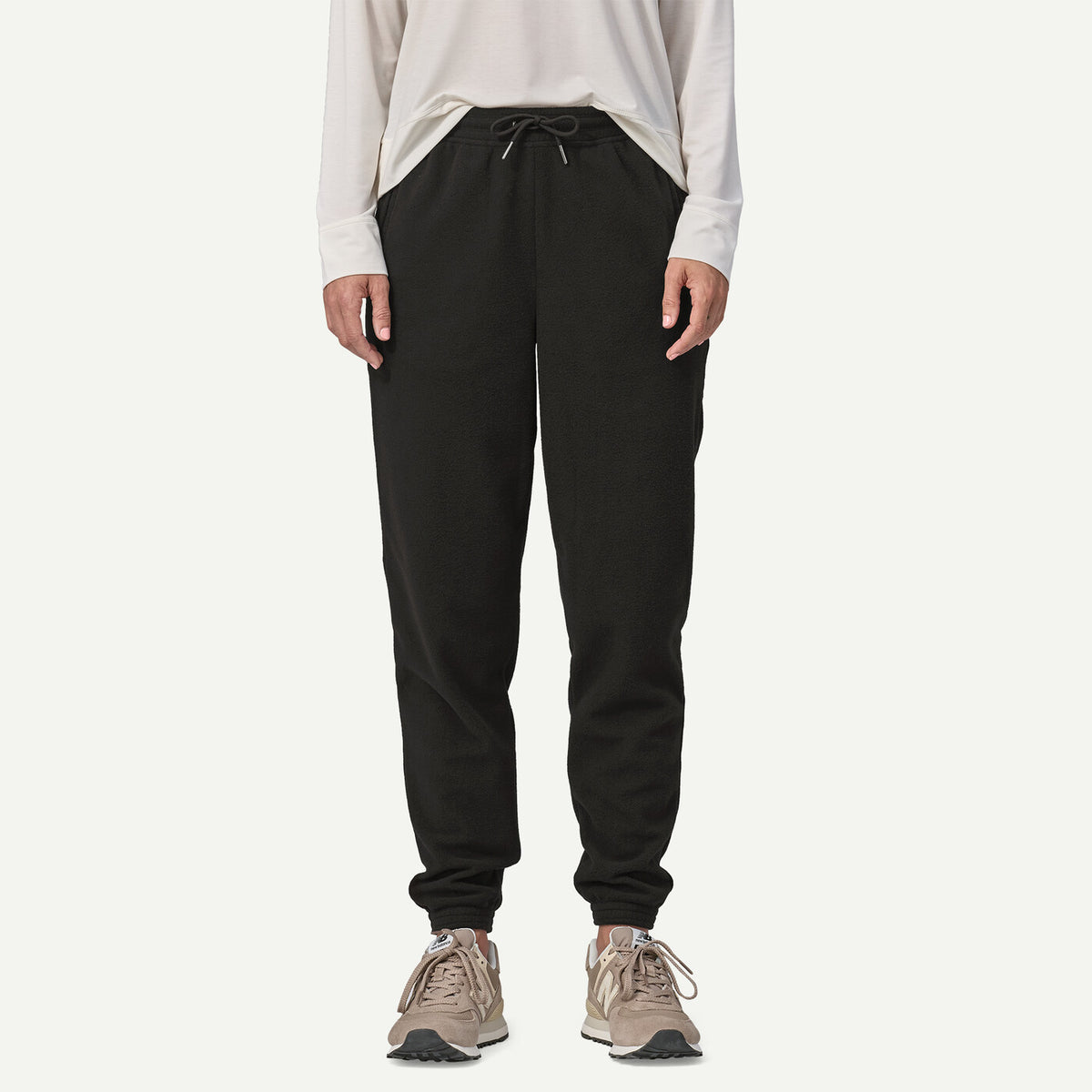A person wearing black jogger sweatpants with a drawstring waist, paired with a light beige long-sleeve top and beige sneakers, shown against a plain light gray studio background.