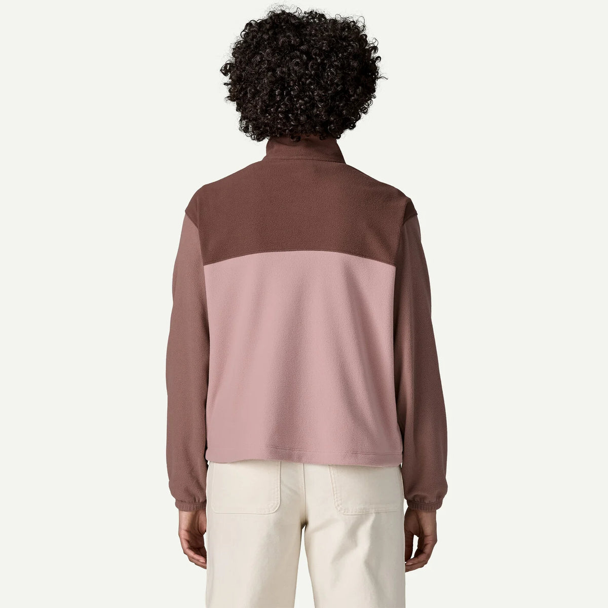 A person shown from the back wearing a color-block fleece pullover with a dark brown top section and sleeves, and a dusty pink lower back panel, paired with cream pants, against a light off-white background.