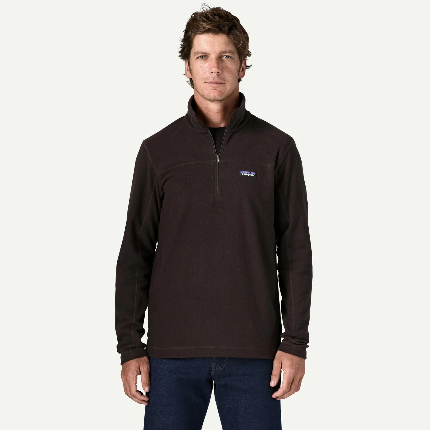 A man wearing a dark brown Patagonia quarter-zip fleece pullover with a small blue and white logo on the left chest, paired with dark blue jeans, against a solid off-white background.