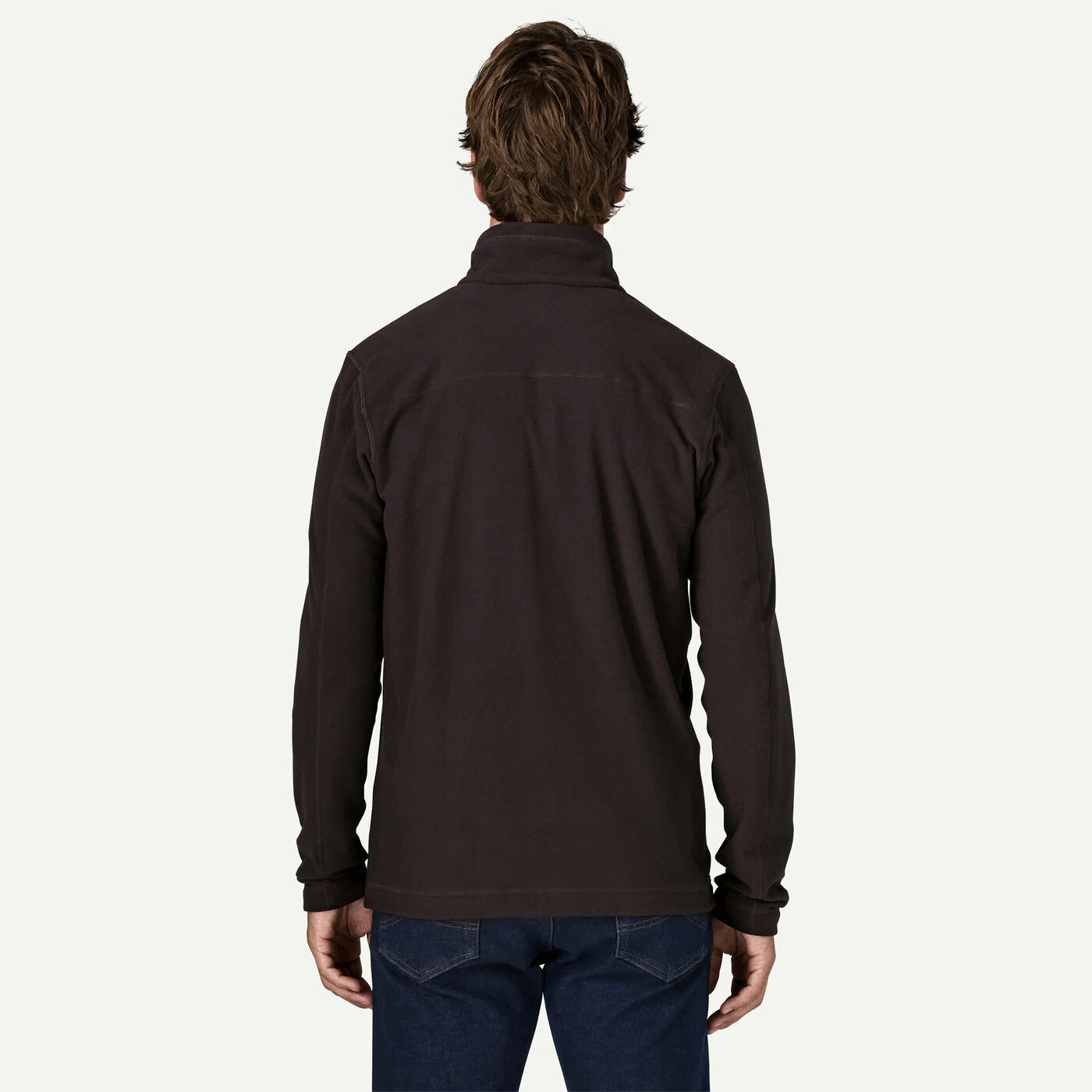 A man viewed from the back wearing a dark brown quarter-zip fleece jacket and blue jeans, set against a plain off-white studio background.