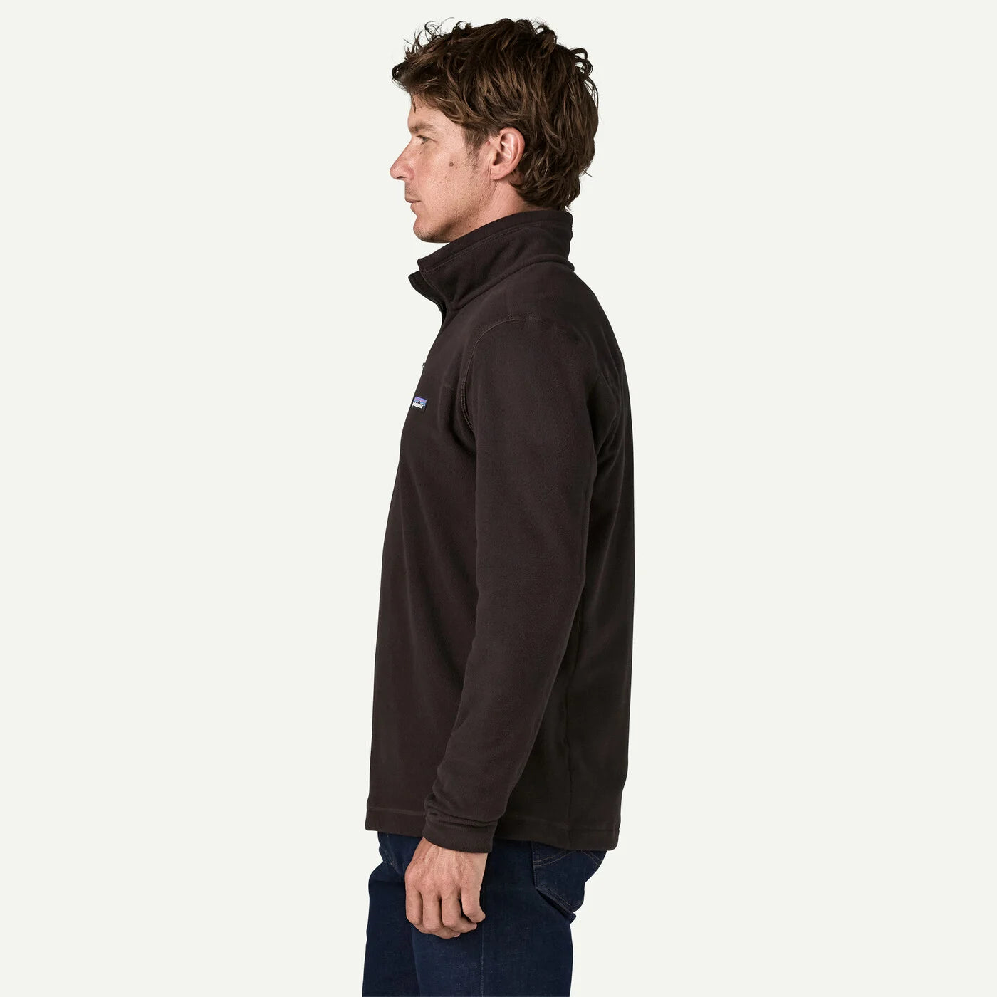 A man in profile wearing a dark brown Patagonia quarter-zip fleece jacket and blue jeans, photographed against a clean off-white studio background.