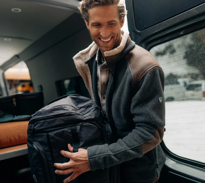 a model wearing the kuhl alpenwurx jacket in steel, moving a bag out of a car