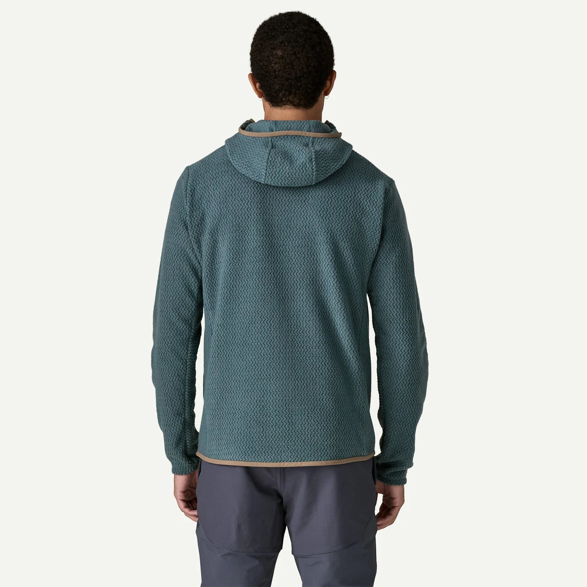 A man viewed from behind wearing a textured teal hooded fleece jacket and dark gray pants against a light off-white background.