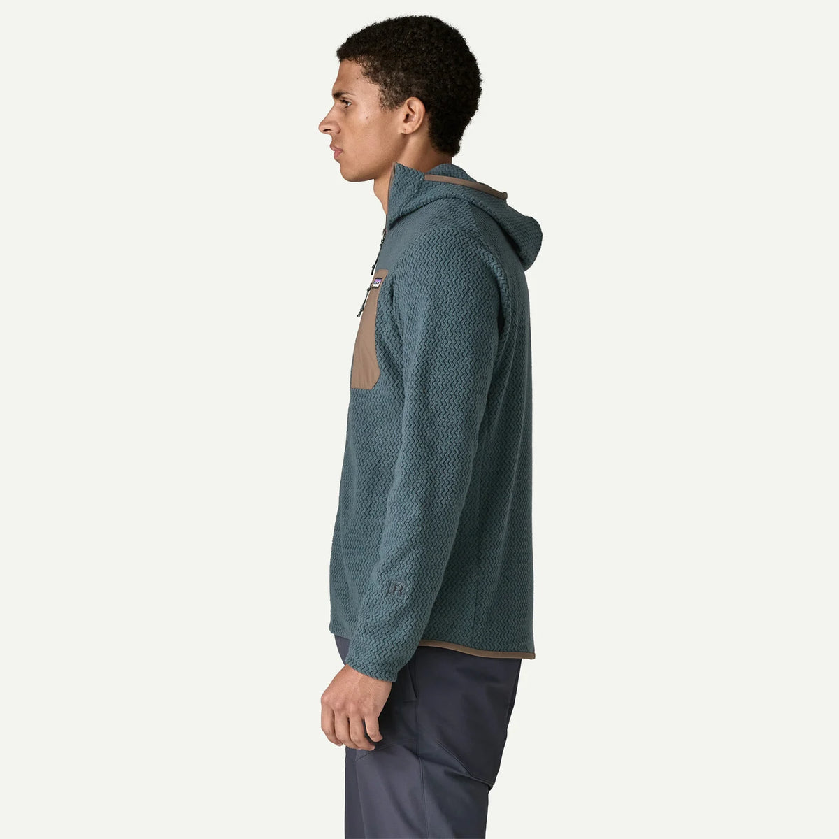 A man in profile wearing a textured slate blue hooded pullover with a tan chest pocket, paired with dark pants, against a solid off-white background.