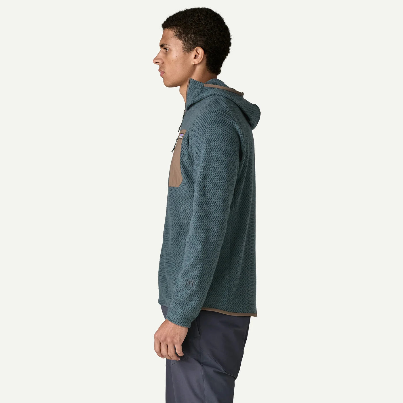A man in profile wearing a textured slate blue hooded pullover with a tan chest pocket, paired with dark pants, against a solid off-white background.