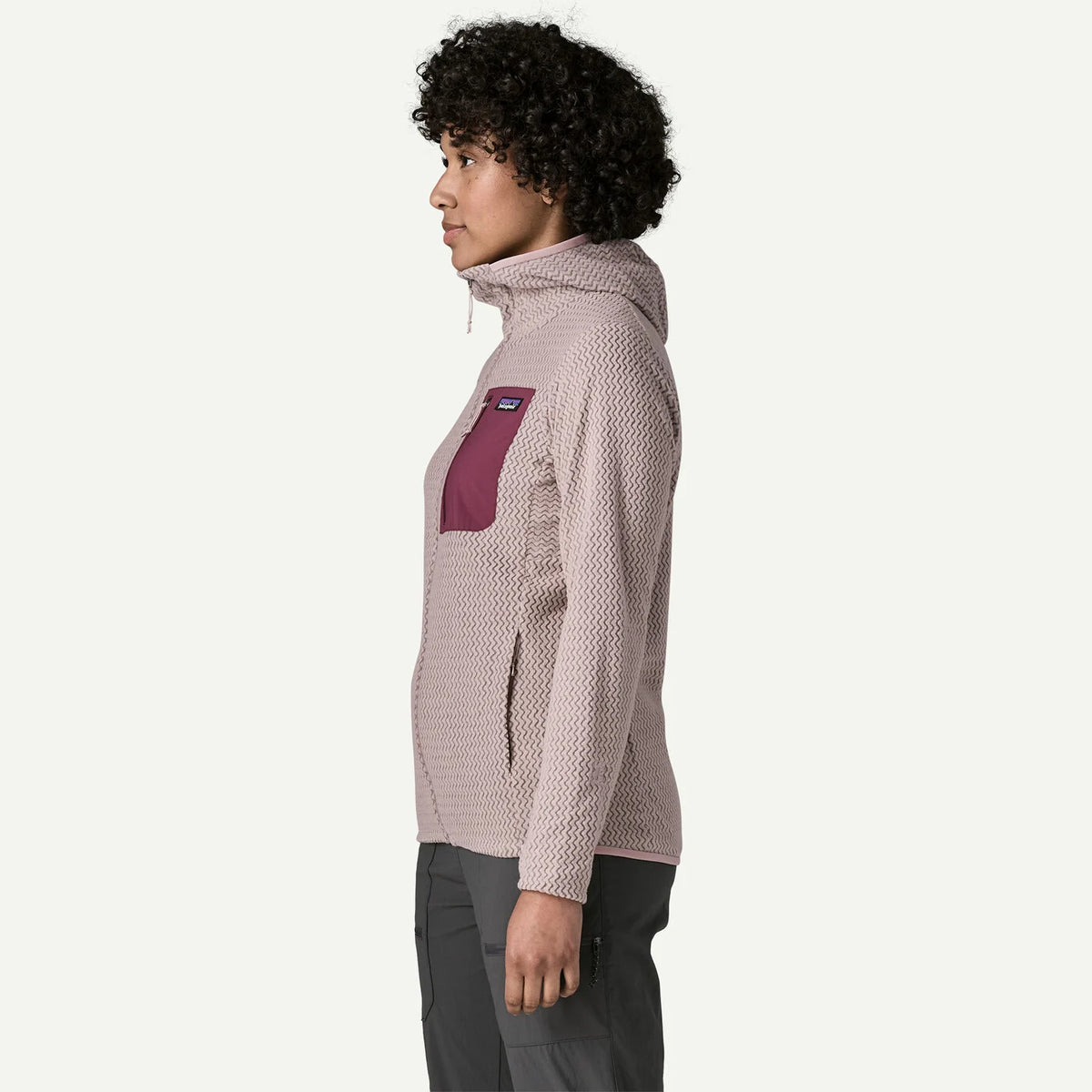 A woman shown in profile wearing a textured zip-up hoodie with a contrasting magenta chest pocket, paired with dark gray pants, against a plain white background.