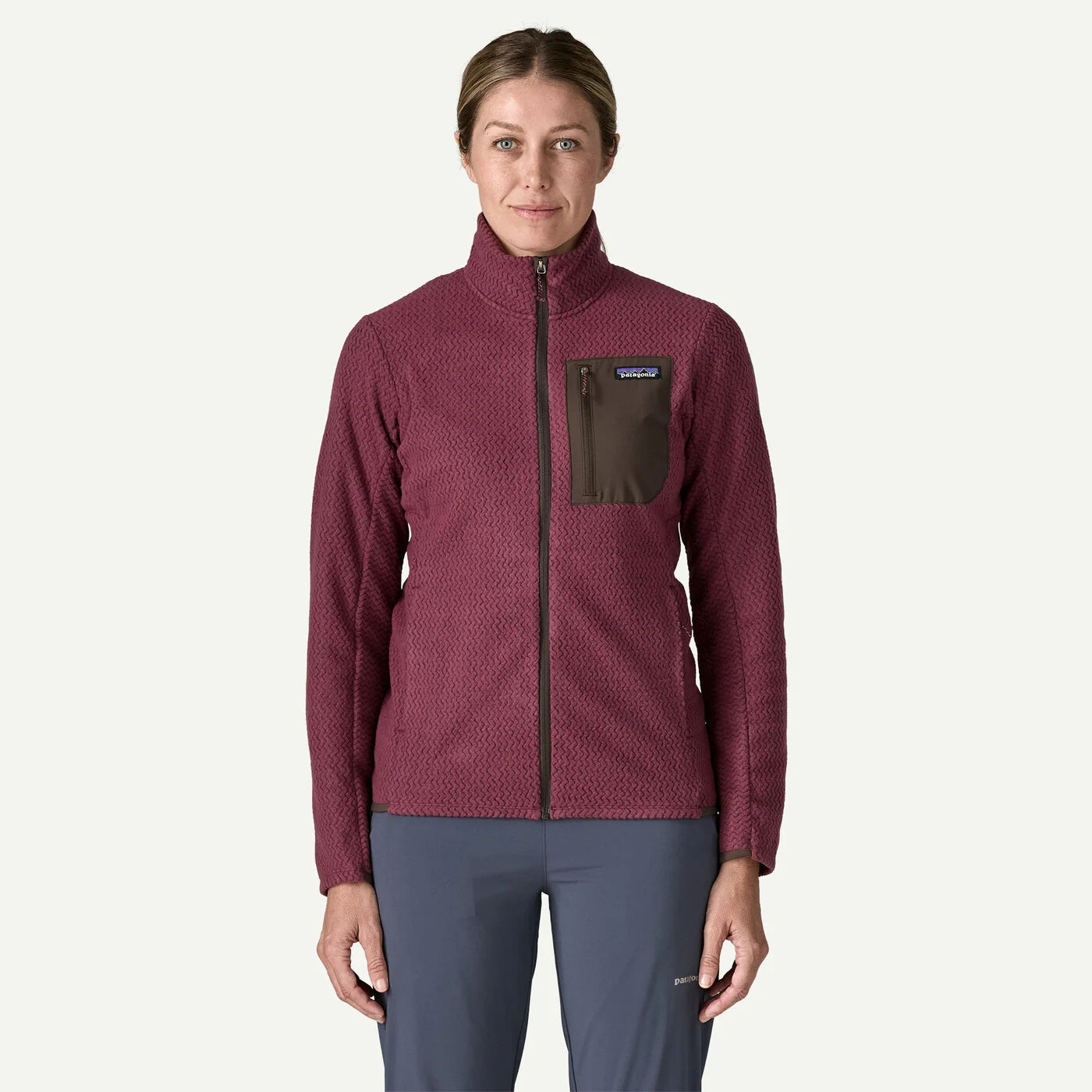 A woman modeling a Patagonia fleece jacket in a textured burgundy color with a dark brown chest pocket, paired with slate blue pants, against a light gray background.