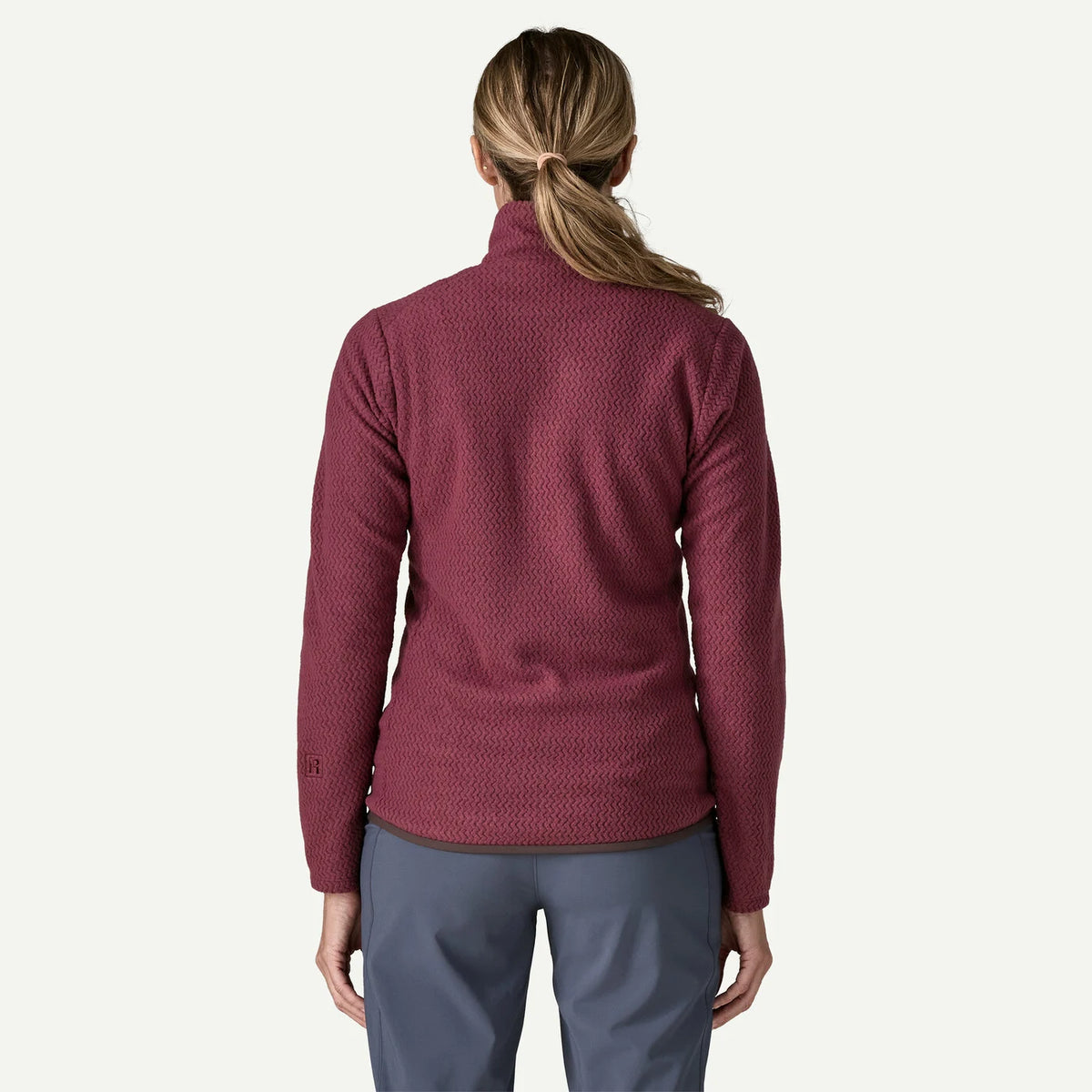 A woman viewed from the back wearing a textured, long-sleeve quarter-zip fleece in a deep burgundy color, paired with slate blue pants, against a plain off-white background.