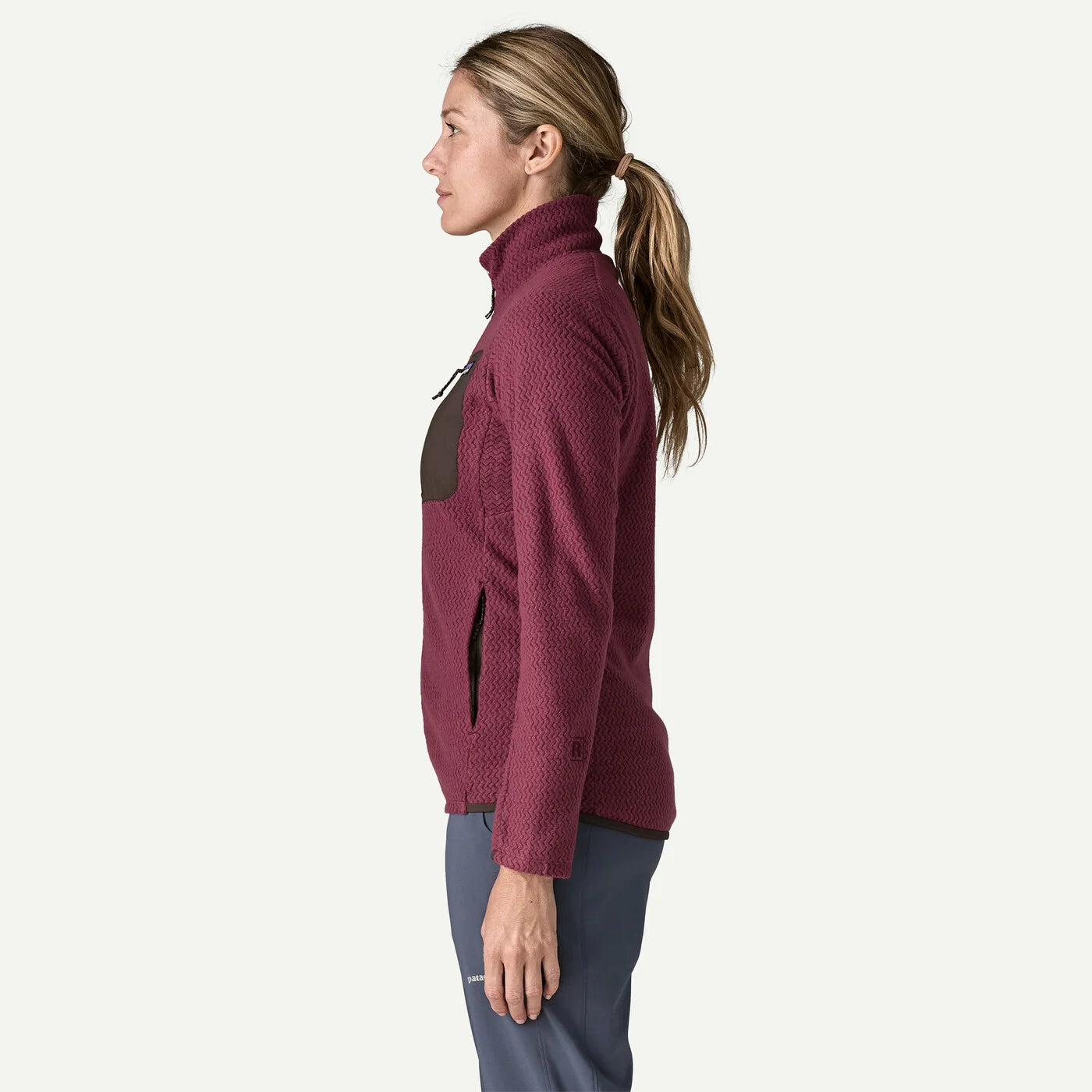 A woman in profile wearing a textured maroon quarter-zip fleece jacket with dark brown contrast panels on the chest and pockets, paired with slate blue pants, against a solid off-white background.