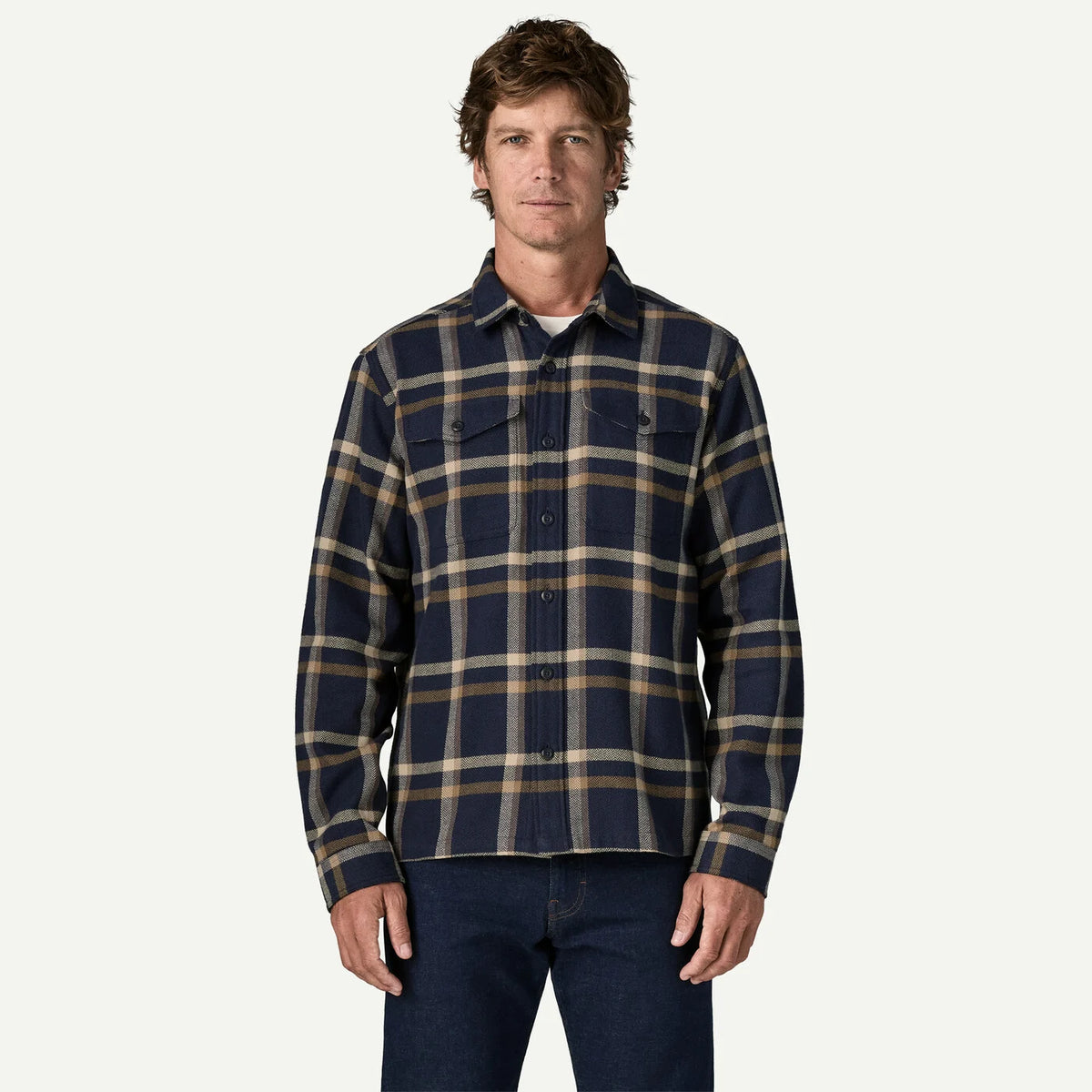 A man wearing a navy blue plaid flannel shirt with tan and beige stripes, paired with dark blue jeans, standing against a light off-white background.