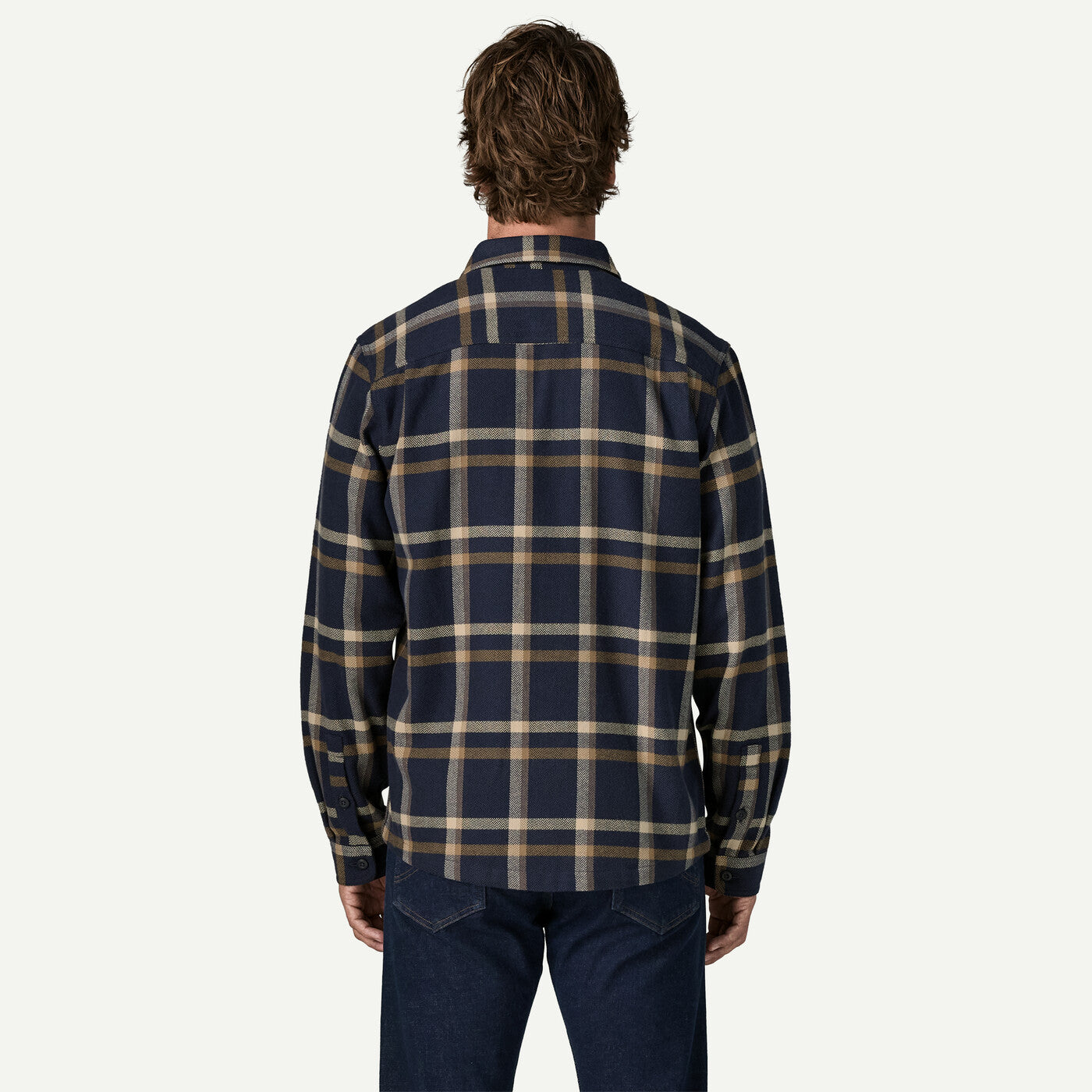 A man viewed from behind wearing a navy blue plaid button-down shirt with tan and beige stripes, paired with dark blue jeans, against a light off-white background.