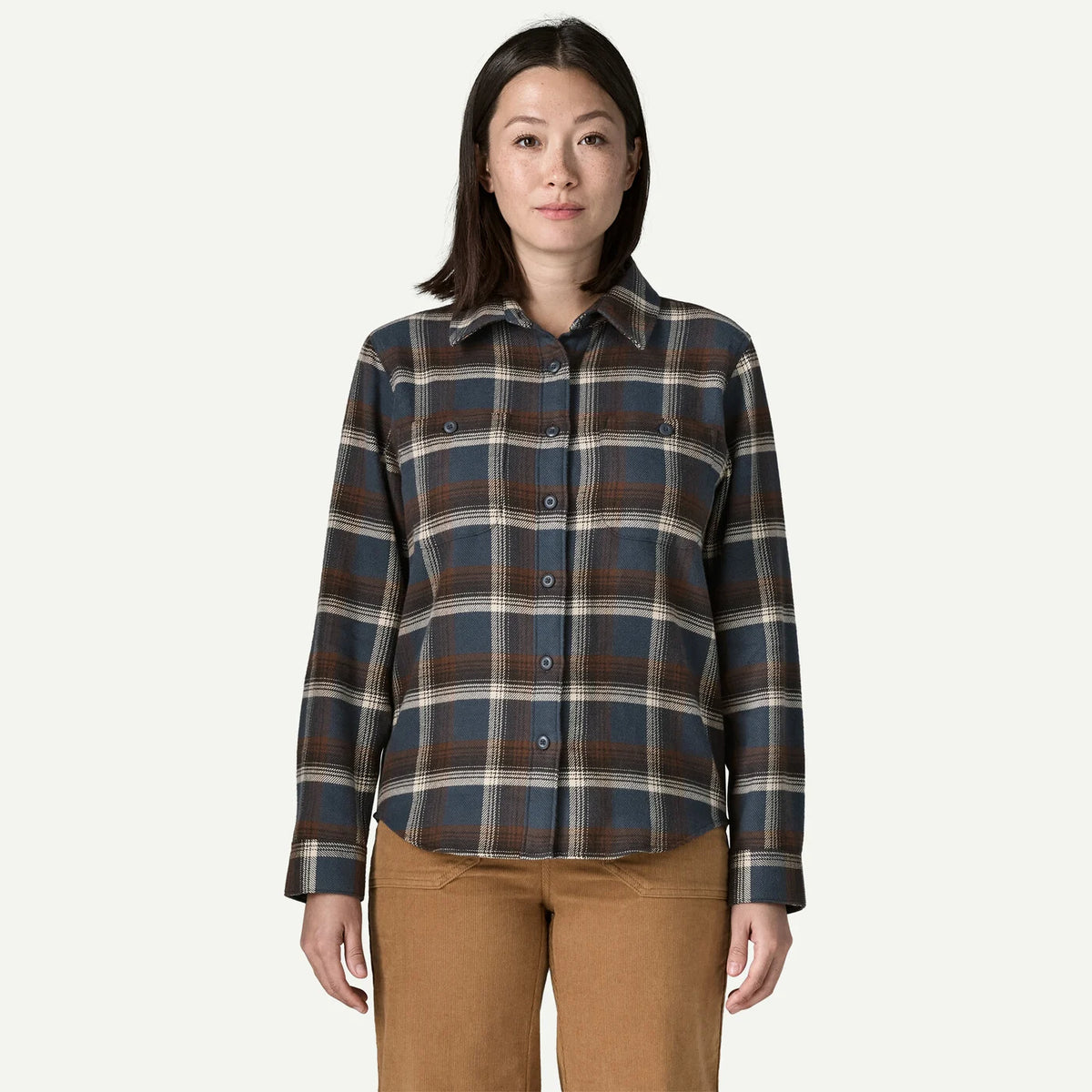 A woman modeling a long-sleeve button-up plaid shirt in dark blue, brown, and beige, paired with tan pants, against a solid off-white background.