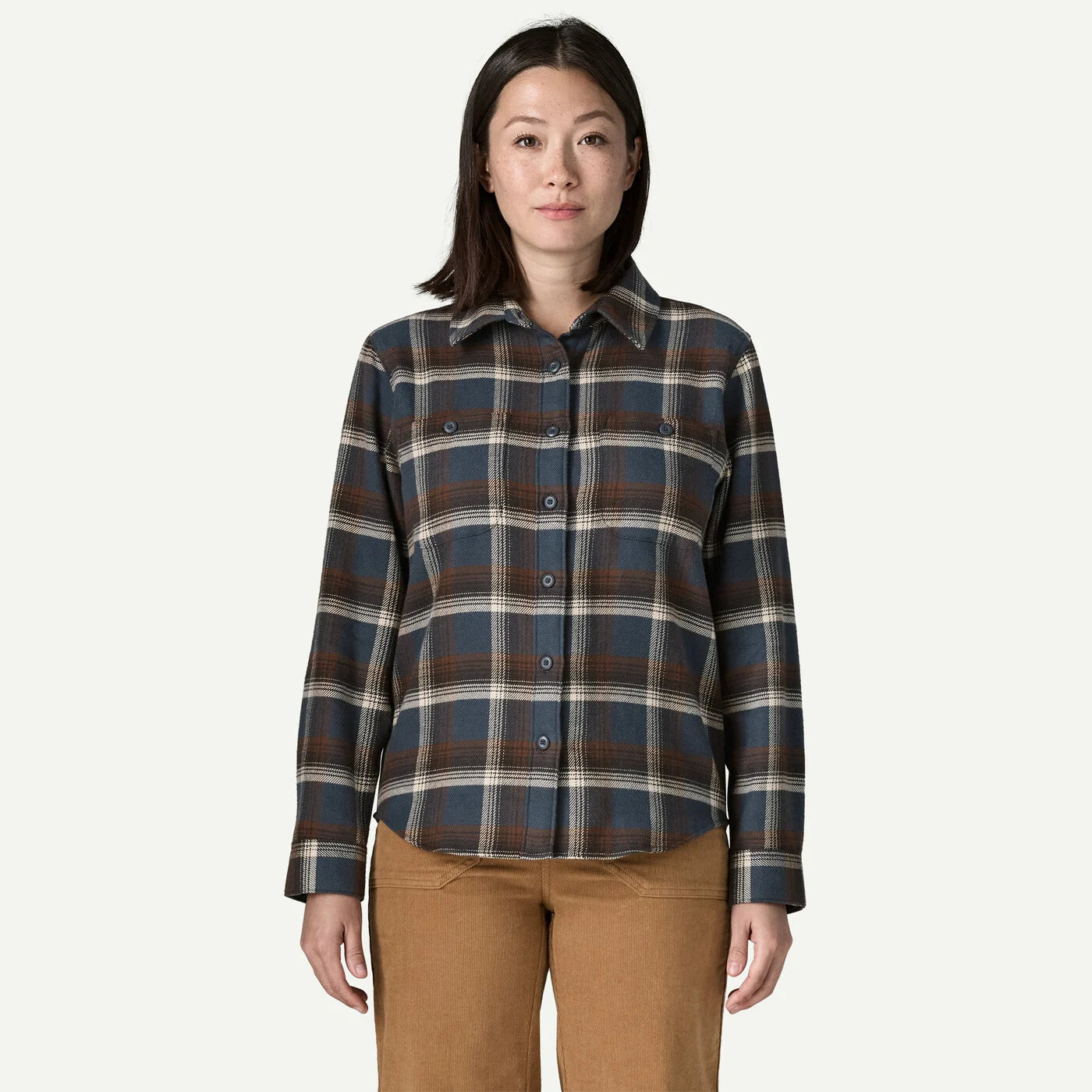 A woman modeling a long-sleeve button-up plaid shirt in dark blue, brown, and beige, paired with tan pants, against a solid off-white background.