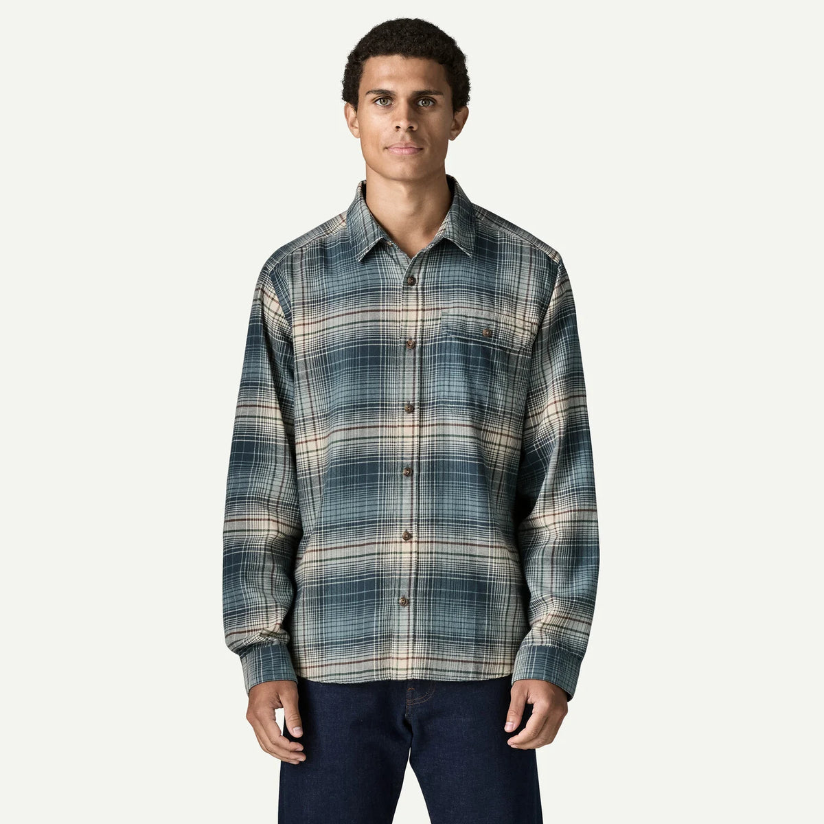A man wearing a long-sleeve button-up plaid shirt in shades of blue, gray, and beige, paired with dark blue jeans, against a light gray background.