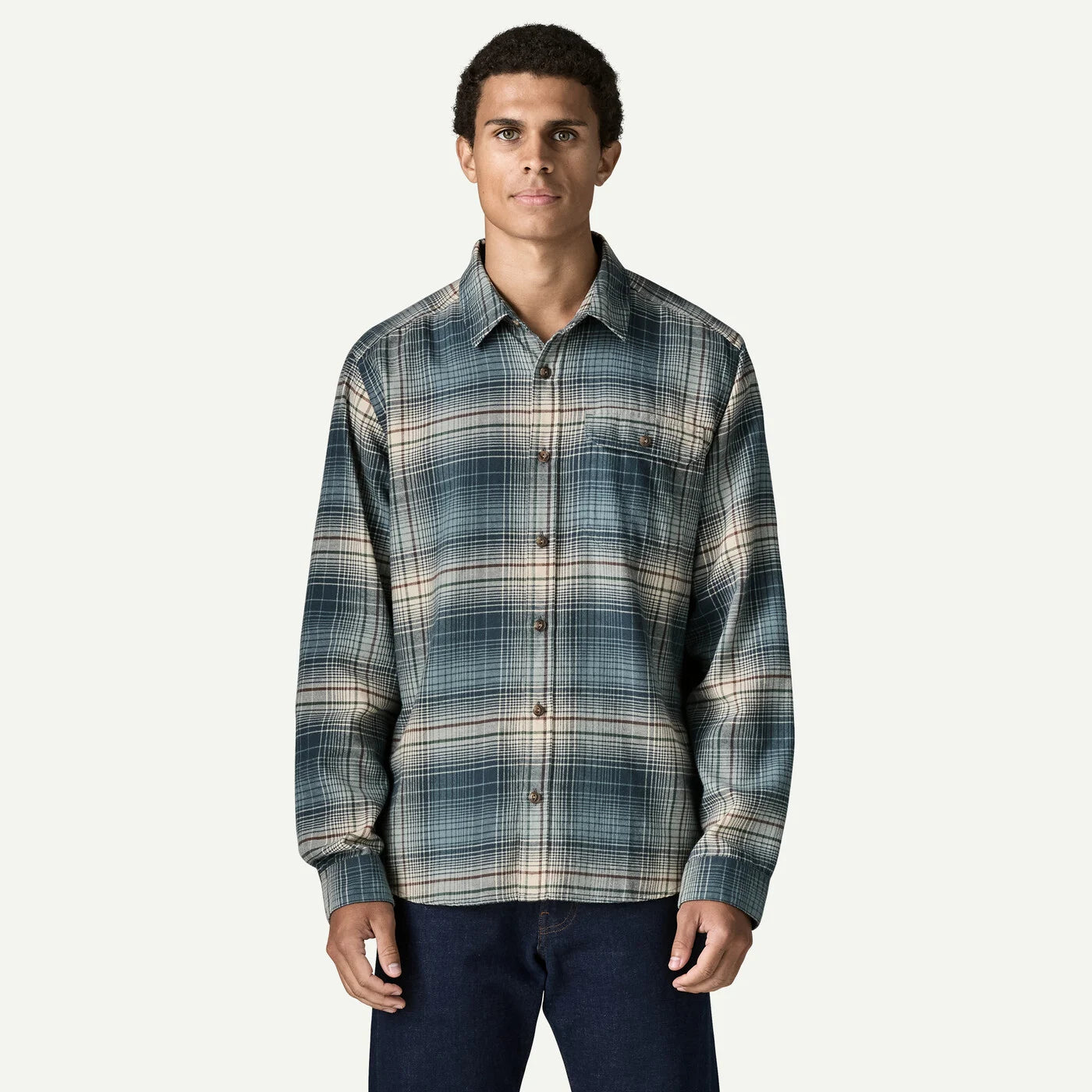 A man wearing a long-sleeve button-up plaid shirt in shades of blue, gray, and beige, paired with dark blue jeans, against a light gray background.