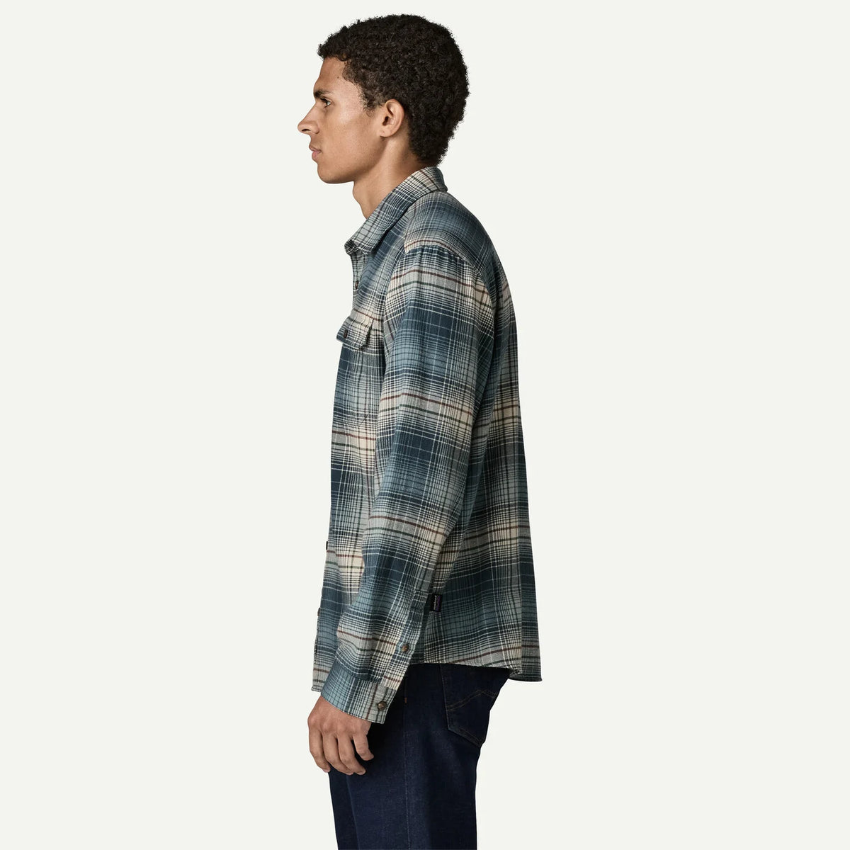 A man shown in profile wearing a long-sleeve plaid button-up shirt in muted blue, gray, and beige tones, paired with dark jeans, against a plain off-white background.