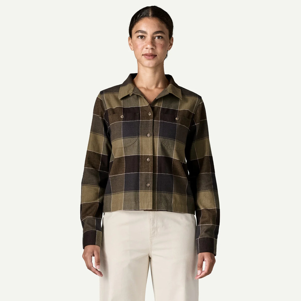 A woman modeling a cropped, long-sleeve plaid button-up shirt in olive green, brown, and black, paired with cream pants, against a solid off-white background.