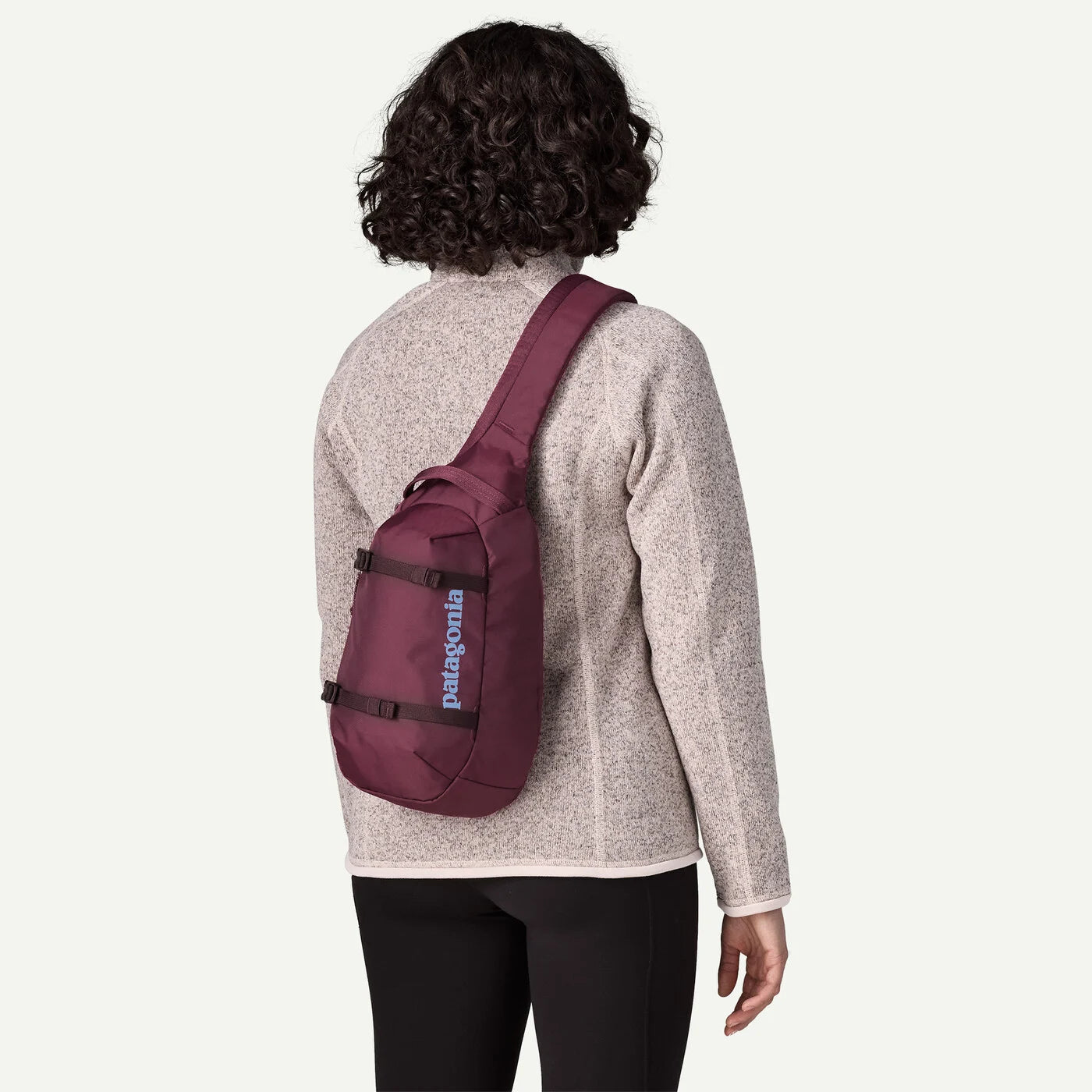 A person shown from the back wearing a Patagonia sling backpack in burgundy against a light gray studio background.
