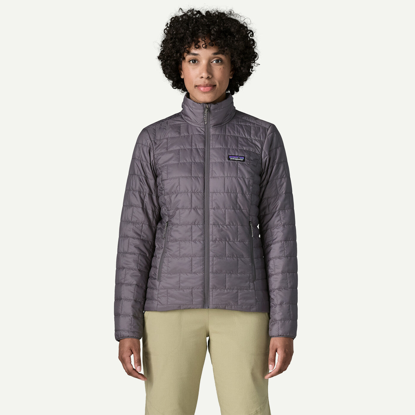 A woman wearing a Patagonia quilted zip-up jacket in a muted grayish purple color, paired with light khaki pants, standing against a solid off-white background.