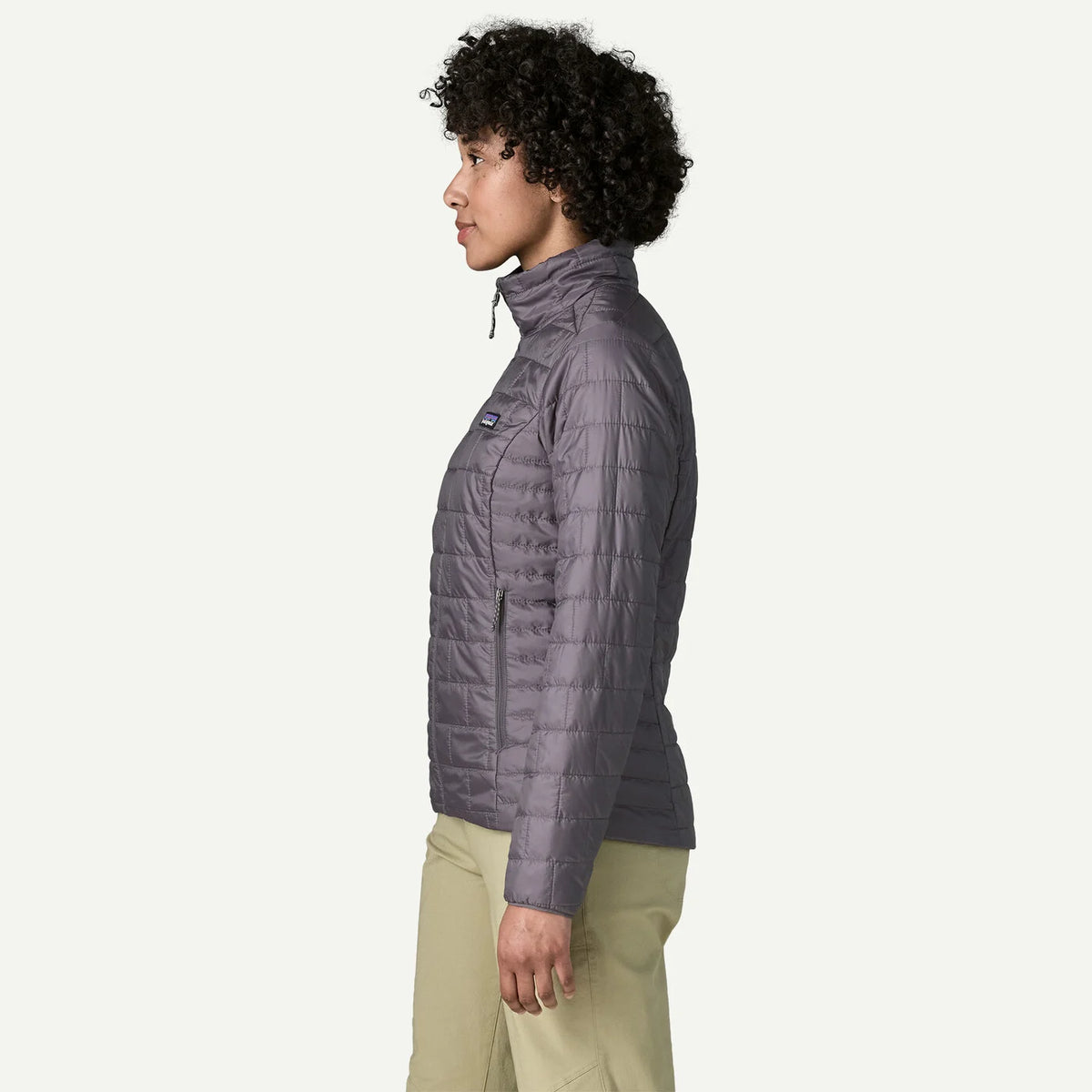 A woman shown in profile wearing a quilted puffer jacket in a muted grayish purple tone, paired with light khaki pants, against a clean off-white studio background.