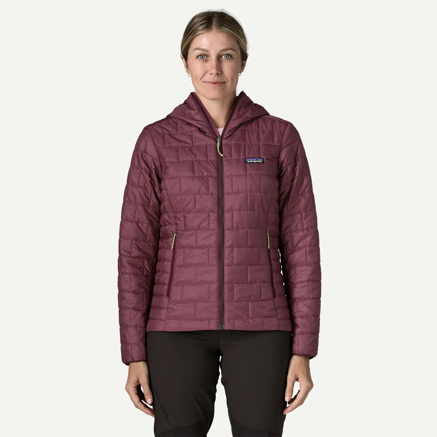 A woman modeling a Patagonia hooded quilted jacket in a dusty plum purple color, paired with black pants, against a solid off-white background.
