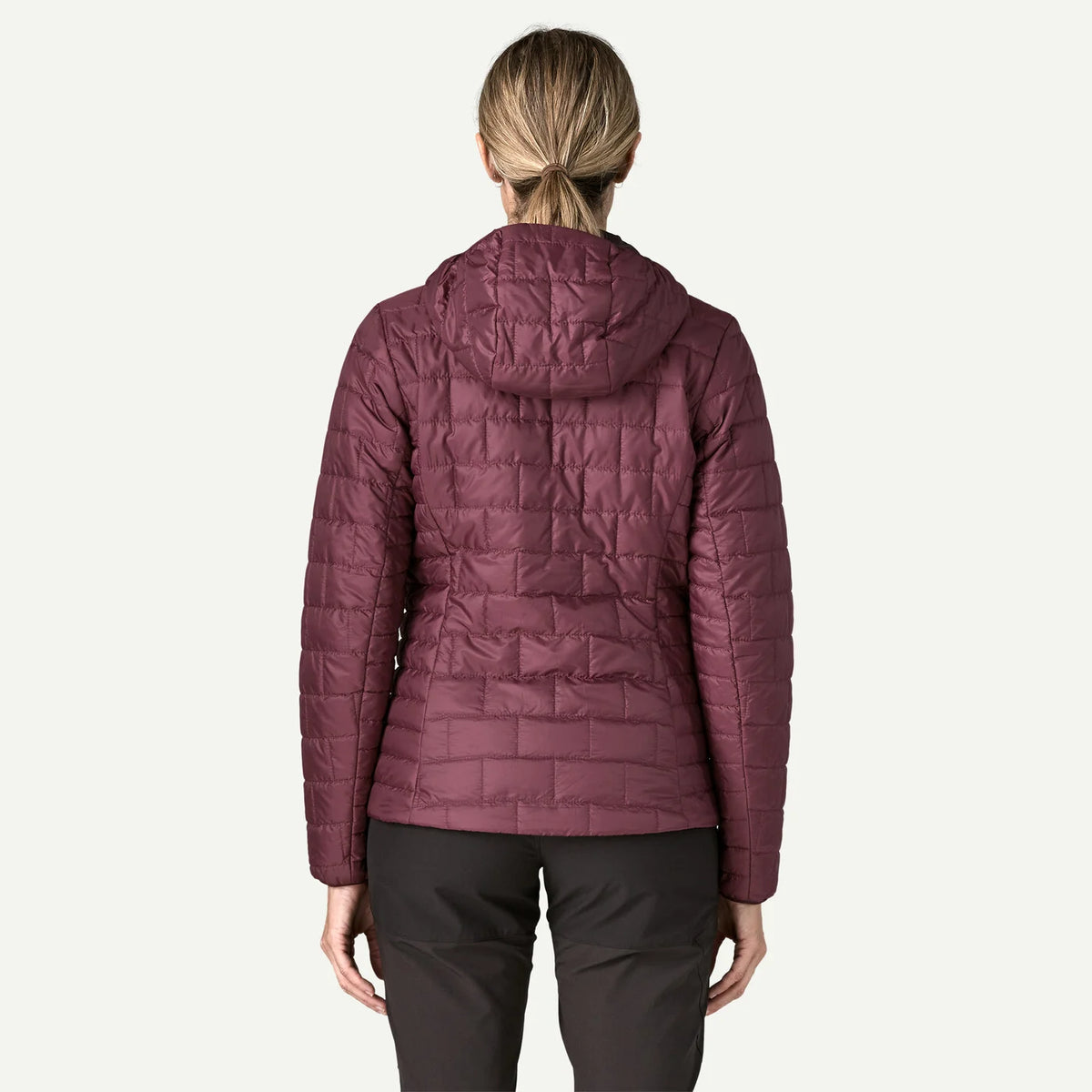A woman viewed from the back wearing a quilted puffer jacket in a muted plum-burgundy color, paired with dark pants, against a plain light background.