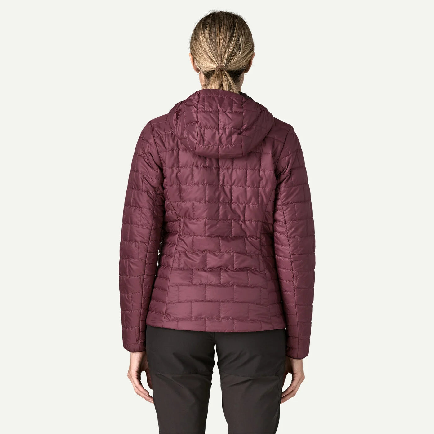 A woman viewed from the back wearing a quilted puffer jacket in a muted plum-burgundy color, paired with dark pants, against a plain light background.