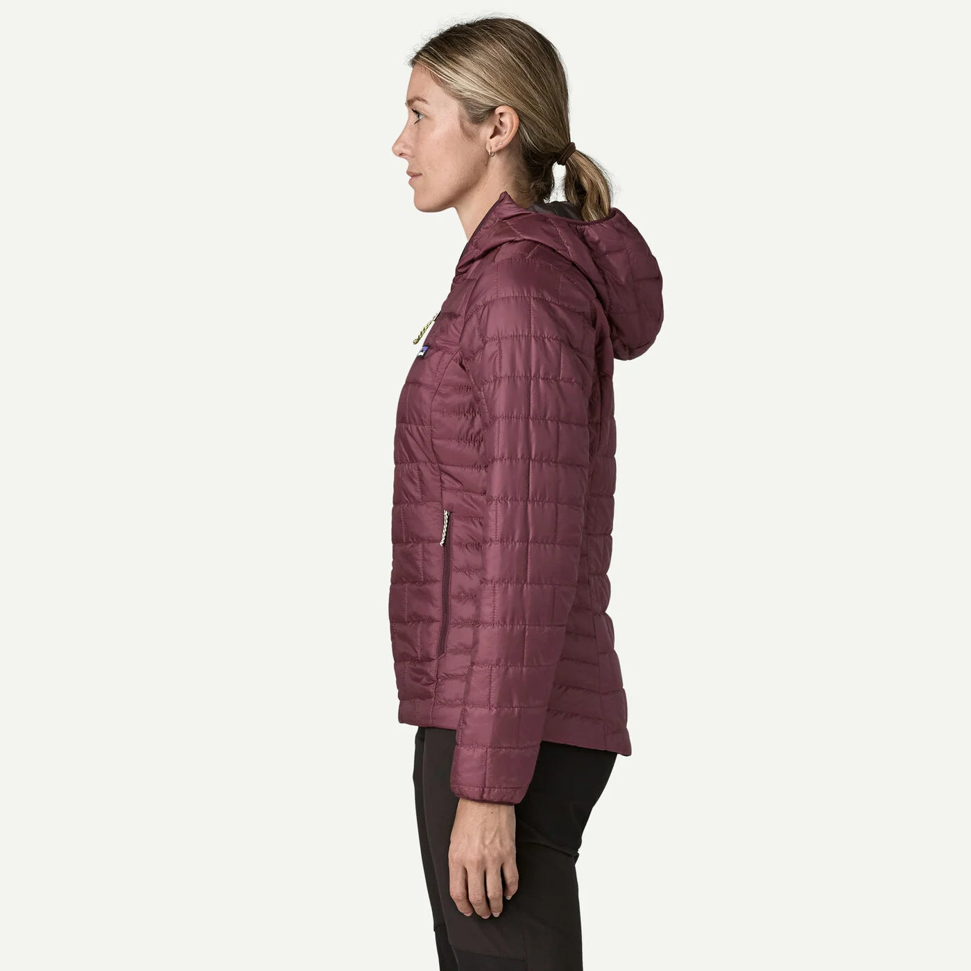 A woman shown in profile wearing a quilted hooded jacket in a muted plum-burgundy color, paired with dark pants, against a plain light gray studio background.