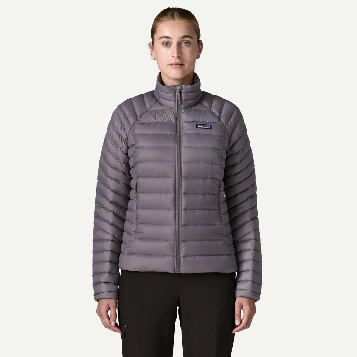 A woman modeling a Patagonia women's quilted puffer jacket in a muted grayish purple color, paired with black pants, against a solid off-white background.