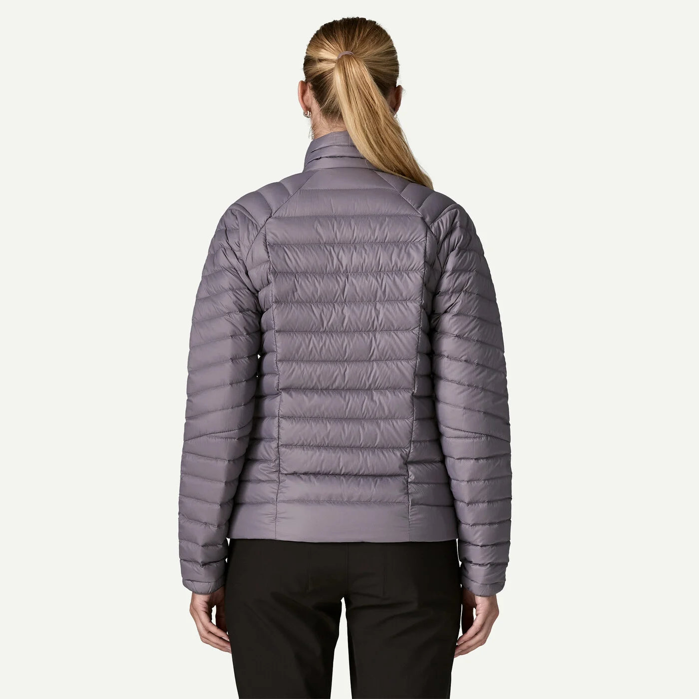 A woman viewed from the back wearing a light purple quilted puffer jacket and black pants, set against a plain off-white background.