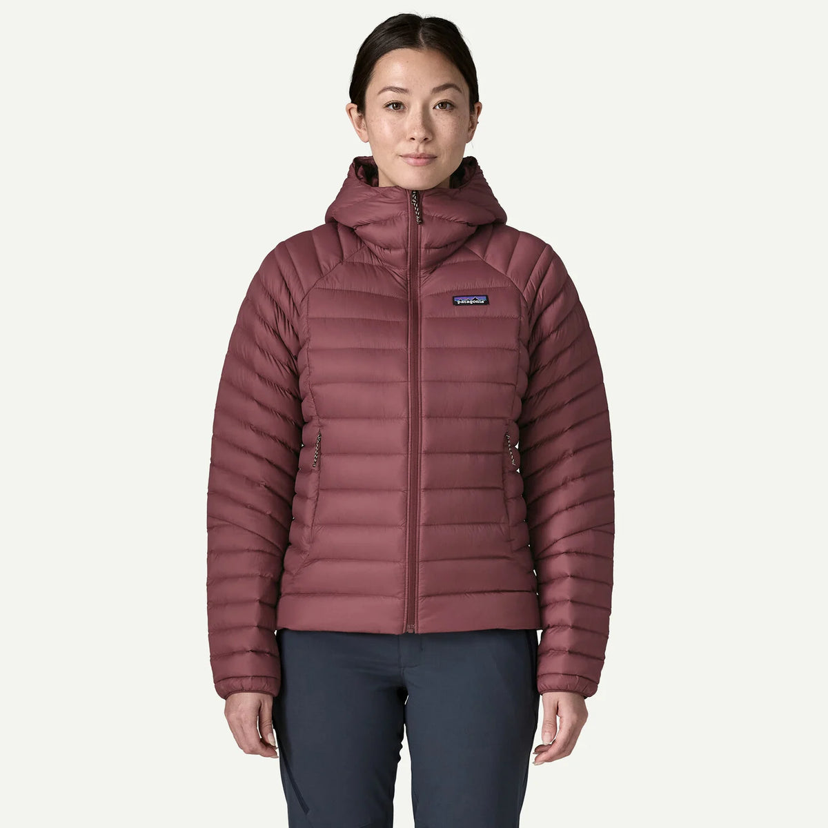 A woman modeling a Patagonia hooded puffer jacket in a dusty rose color, paired with dark gray pants, against a light off-white background.