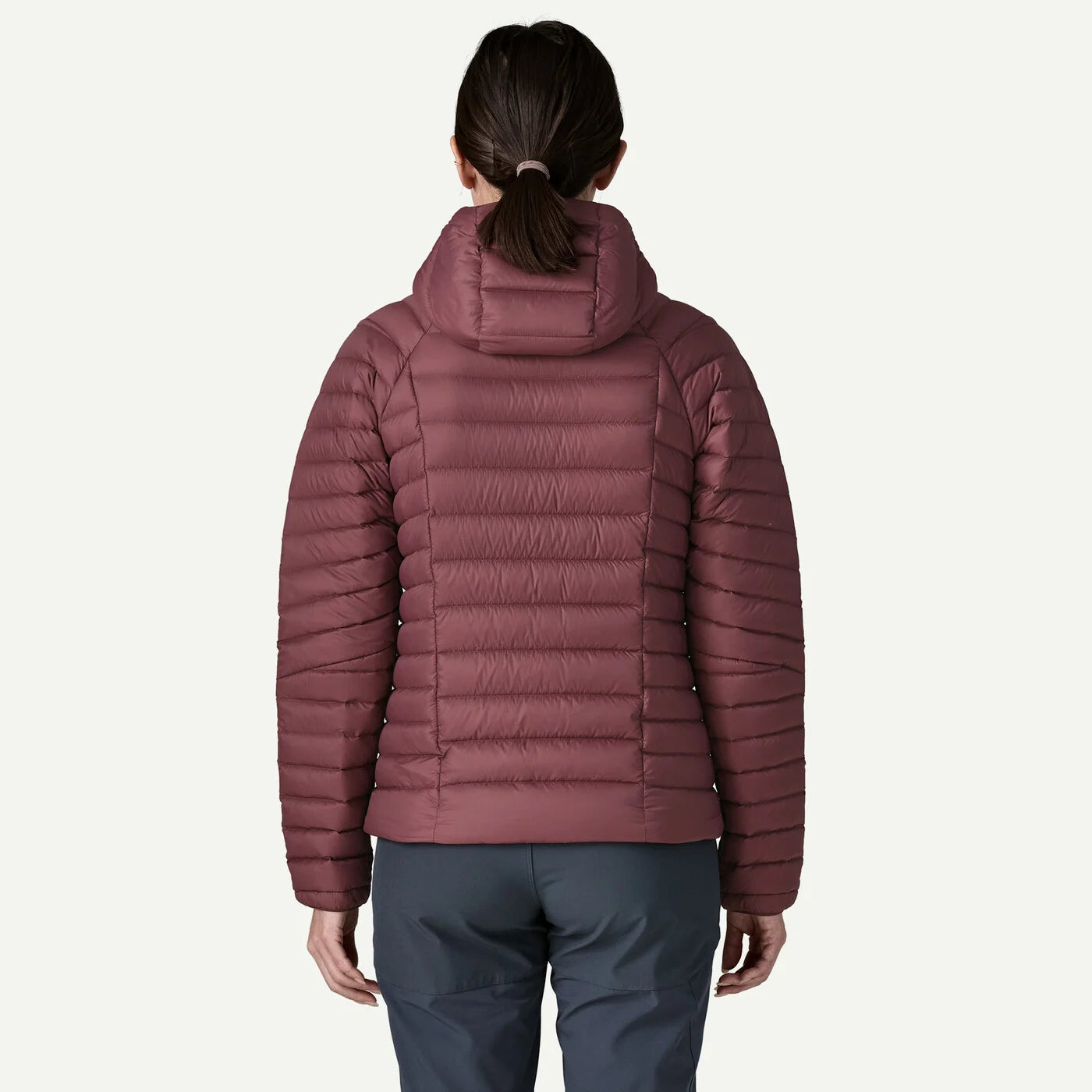A woman viewed from the back wearing a quilted puffer jacket in a muted dusty rose or mauve color, paired with dark gray pants, against a plain off-white studio background.