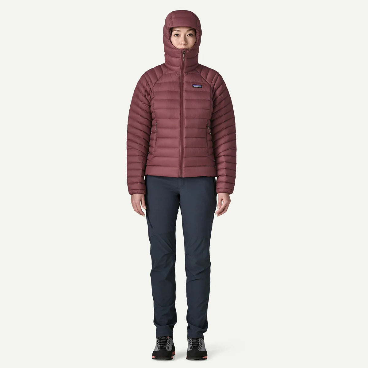A model wearing a Patagonia women's insulated hooded puffer jacket in a dusty rose color, paired with dark navy pants and black hiking boots, standing against a light off-white background.