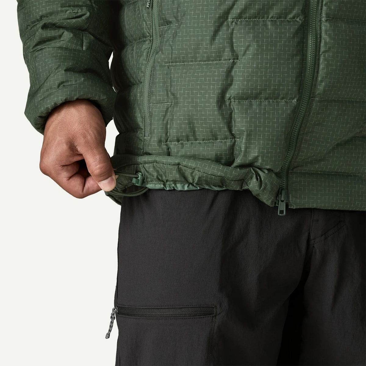Close-up of a person wearing a dark green quilted puffer jacket and black pants, pulling the hem of the jacket down slightly with one hand against a light gray background.